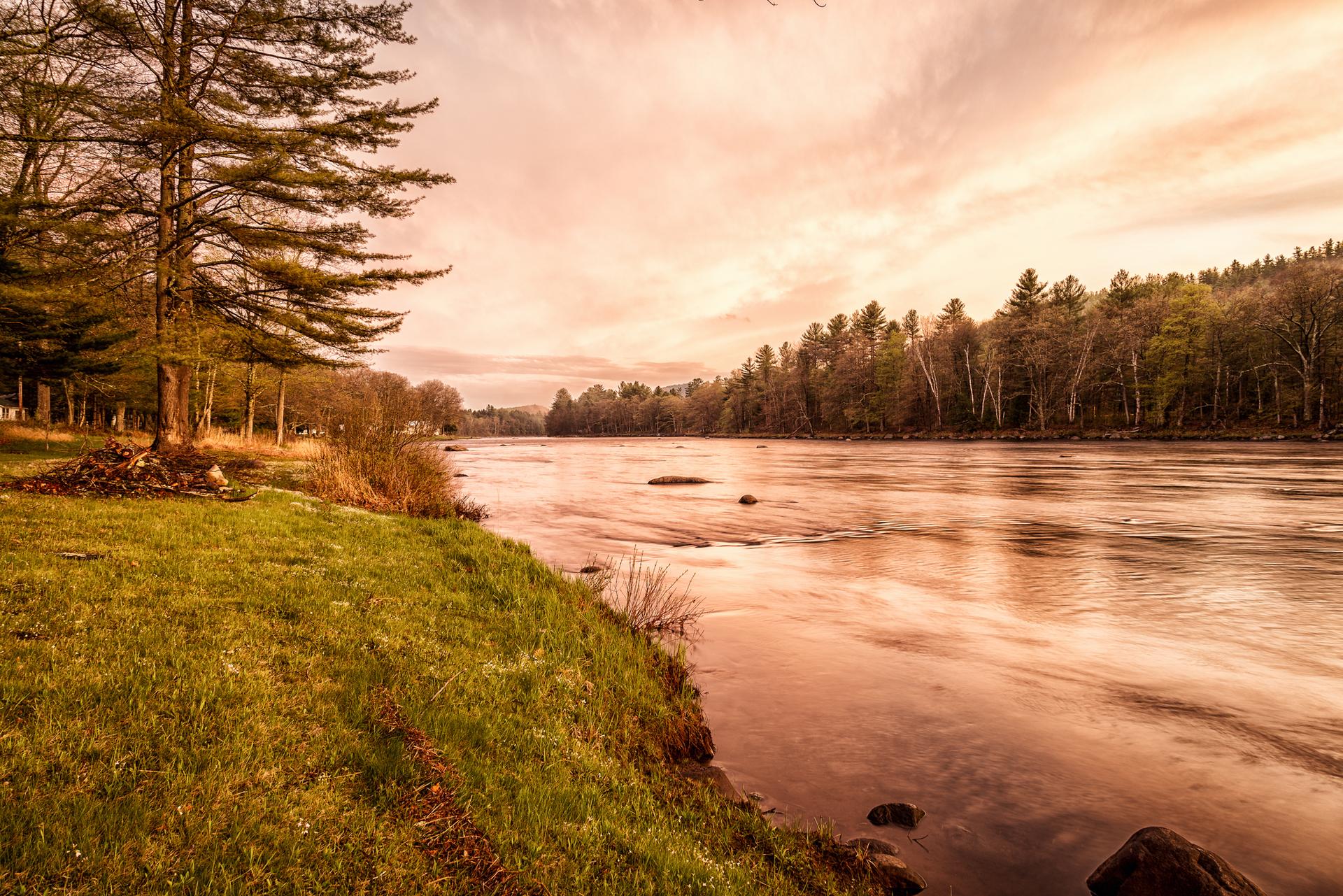 A wide river flows past a grassy bank and forest under a warm, cloudy sky.