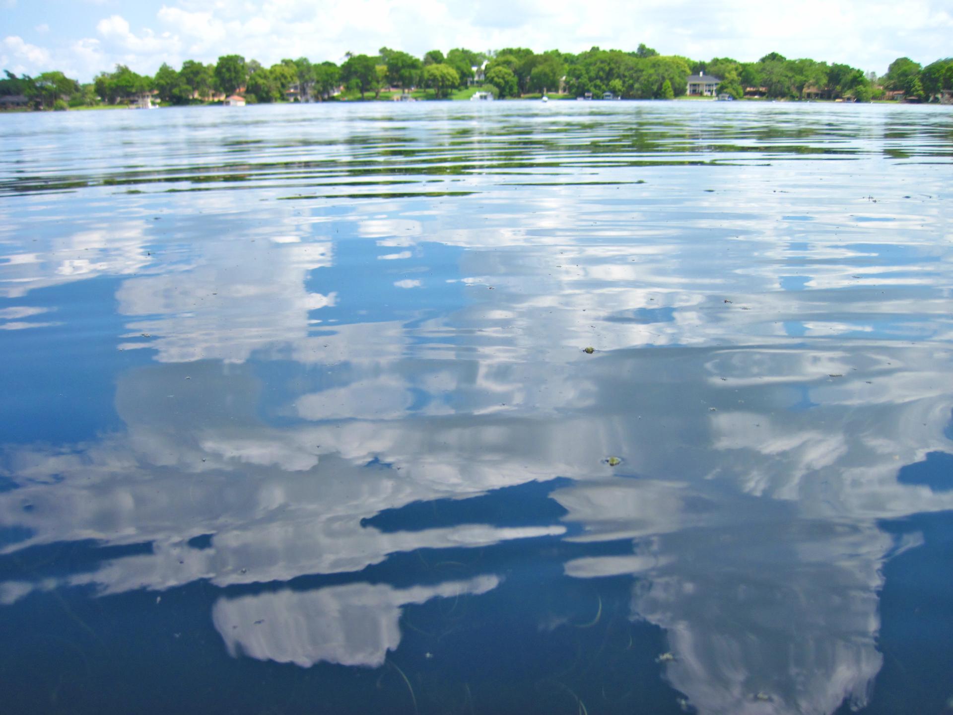Blue lake water reflecting white clouds, with a distant treeline.