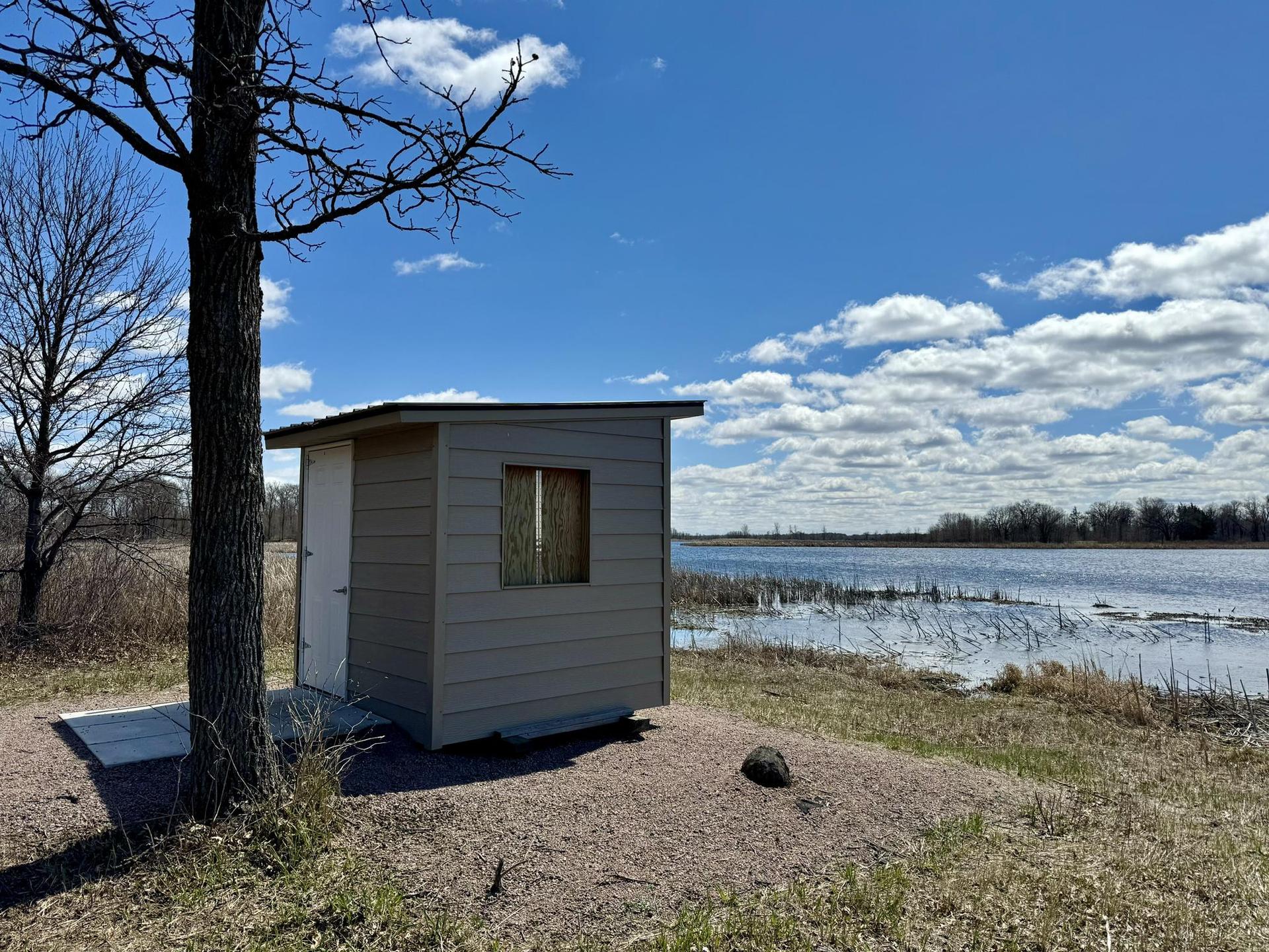 A small gray shed by a tree, overlooking a lake under a blue sky.