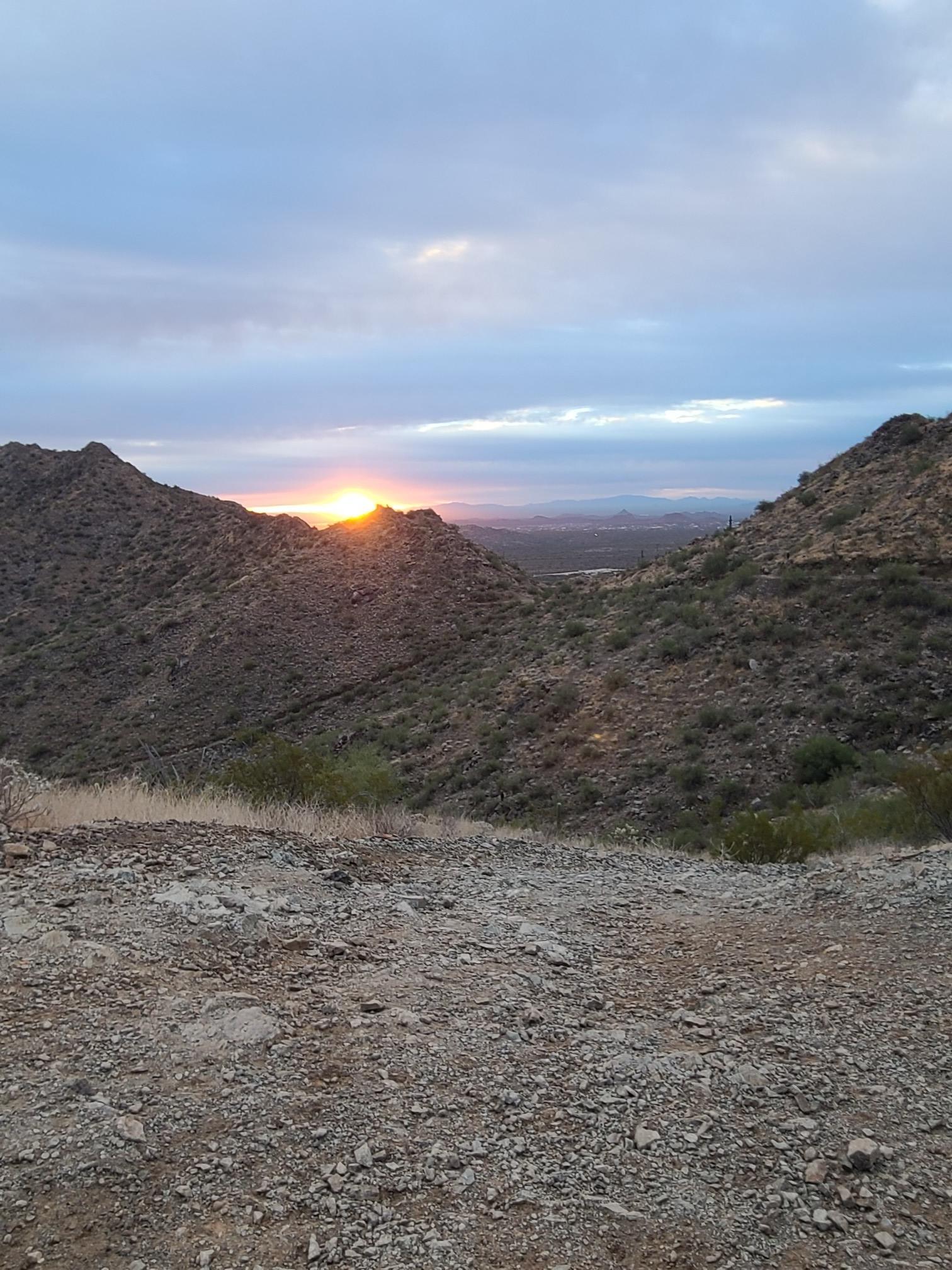 Sunset over rugged desert mountains with a dirt foreground under cloudy sky.