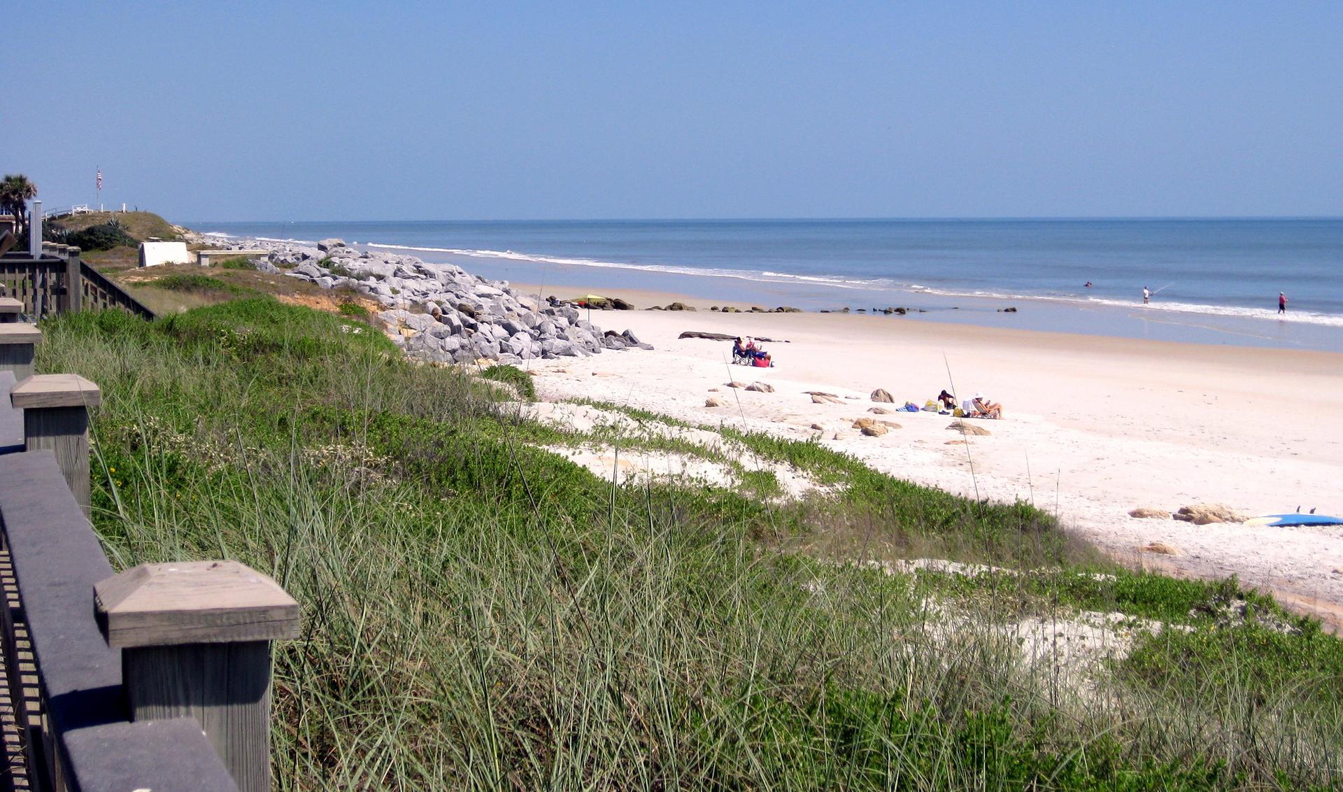 Sunny beach with grassy dunes, ocean, and a wooden boardwalk.
