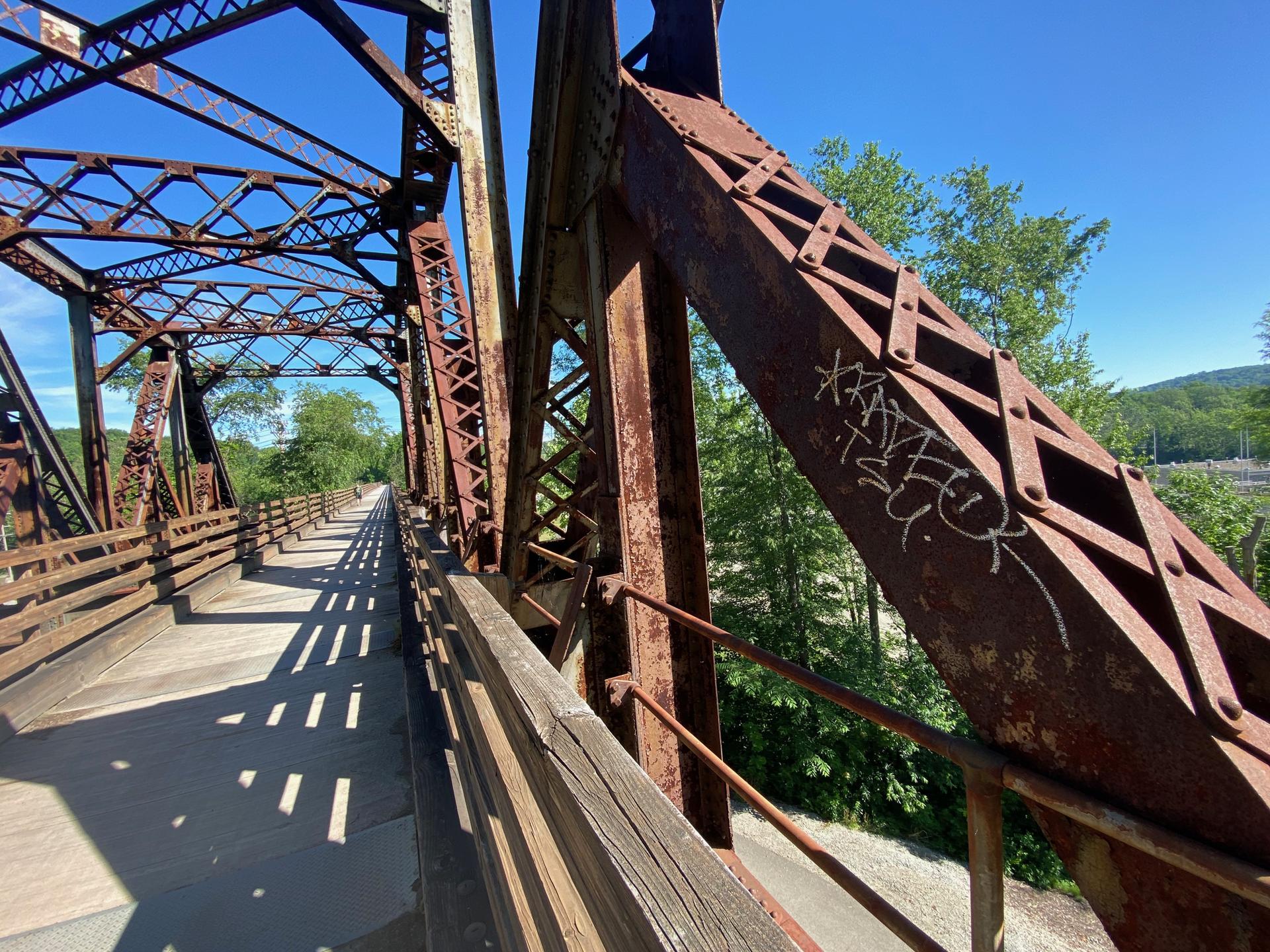 A rusty metal truss bridge with a wooden deck on a sunny day.