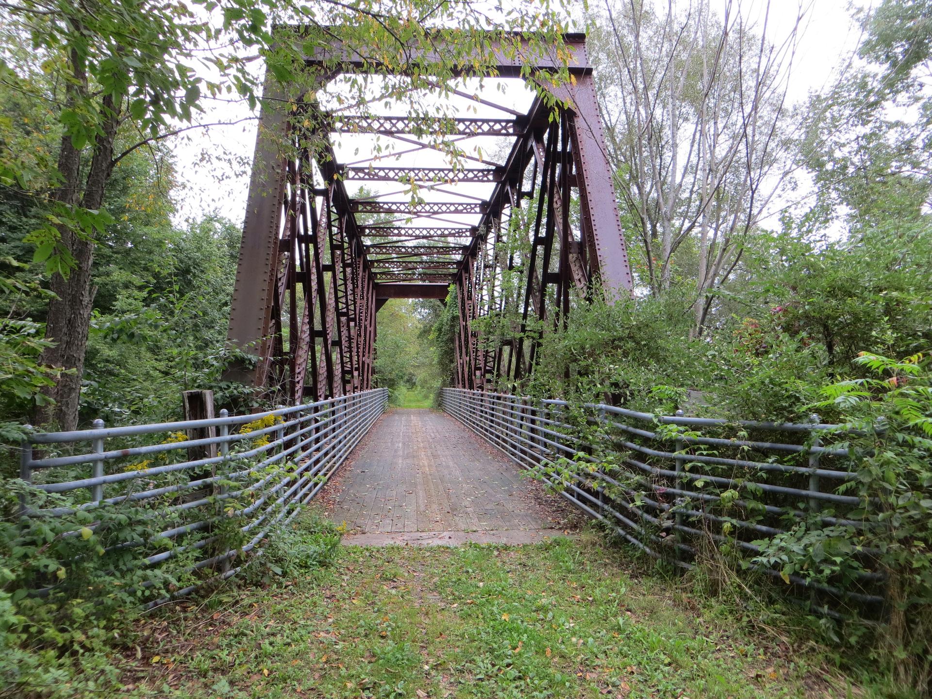 A rusty metal truss bridge spans a dirt path, surrounded by green trees.
