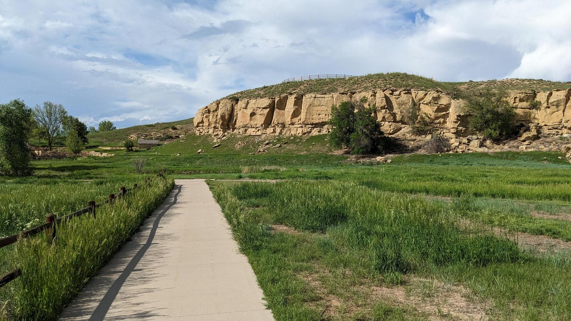 Paved path through green fields towards a large rocky cliff under a cloudy sky.