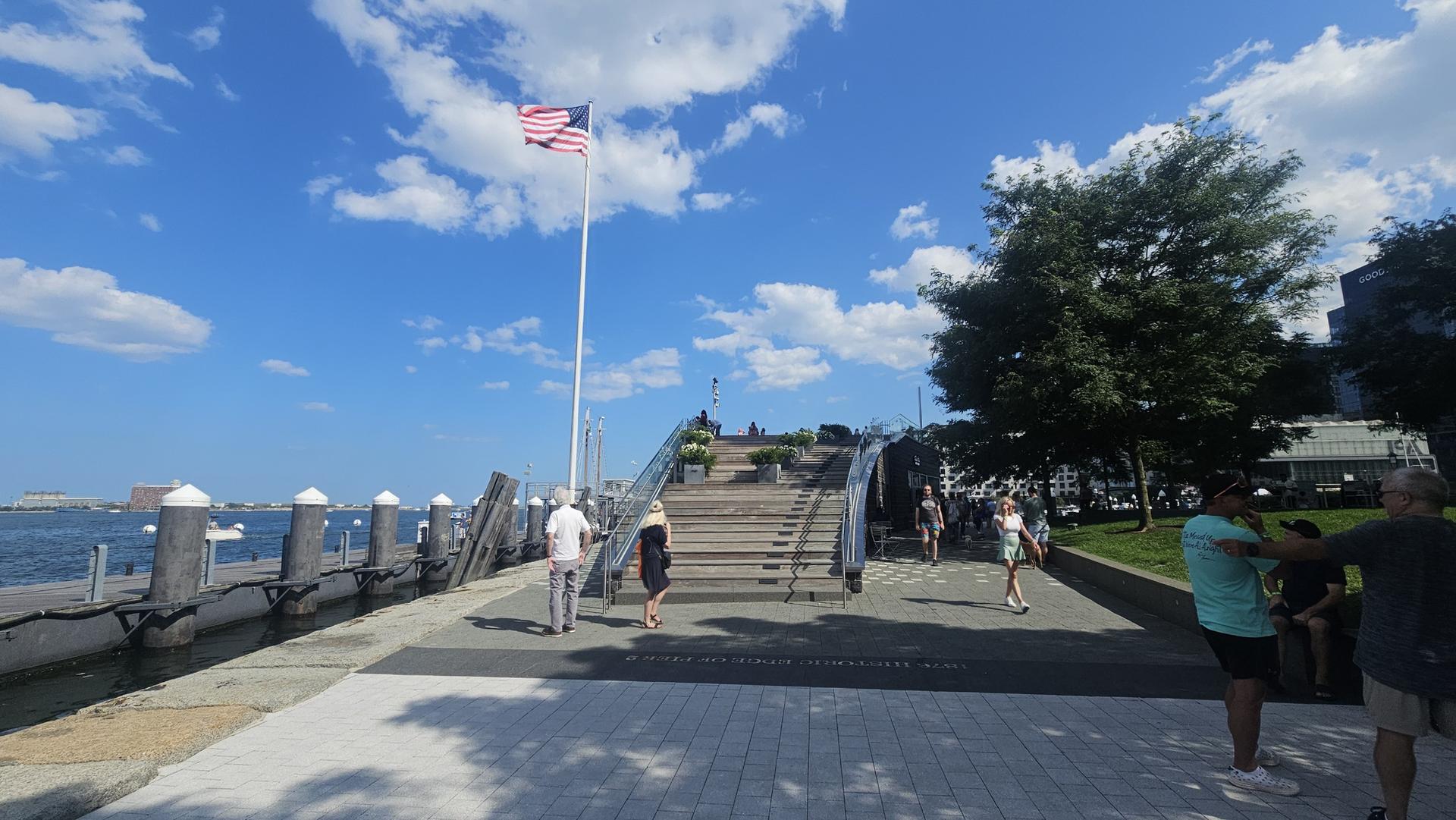People walking on a pier with an American flag flying under a blue sky.