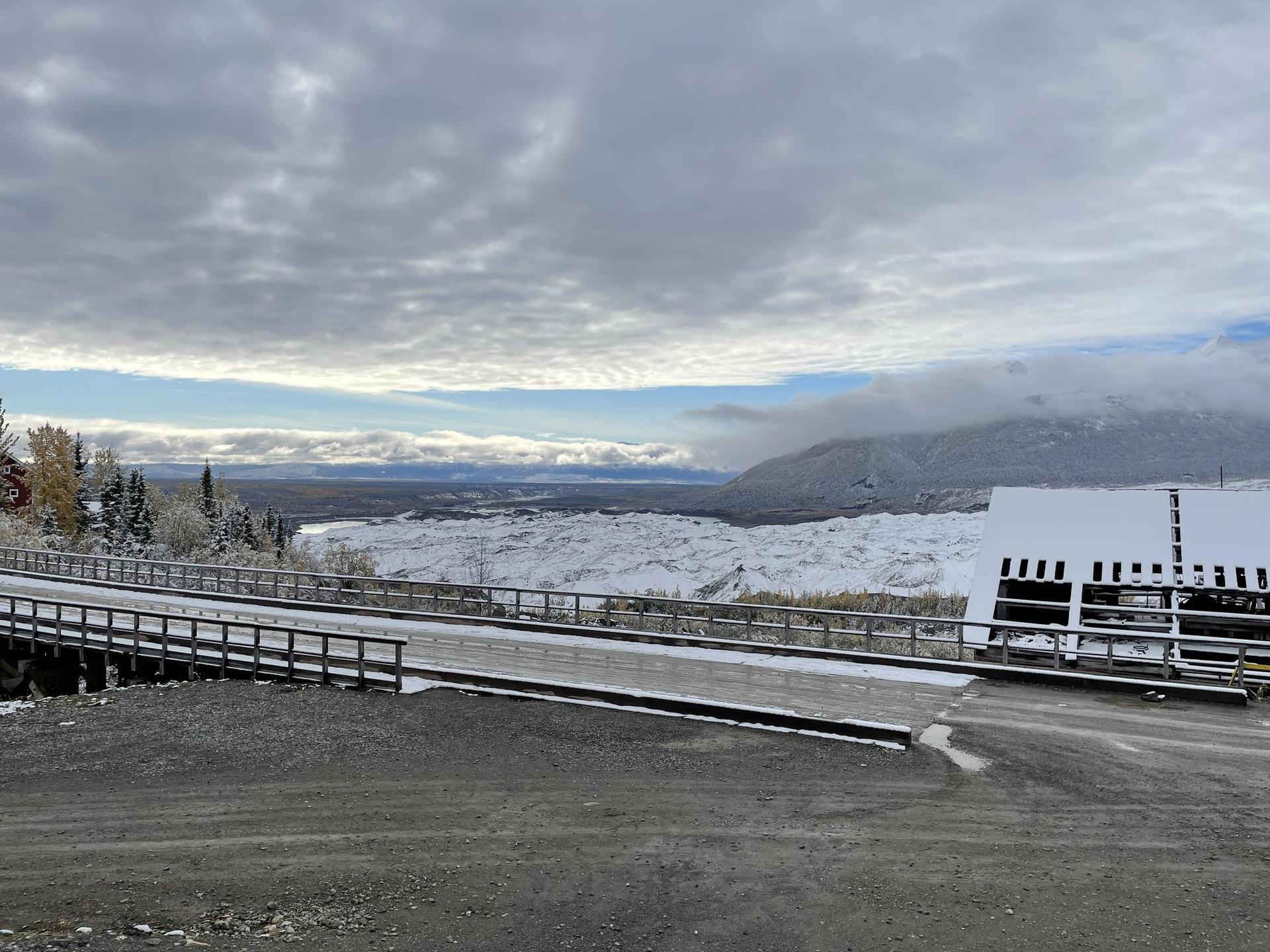 A snowy landscape with a bridge and distant mountains under a cloudy sky.