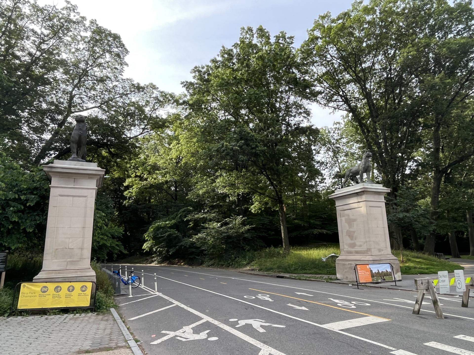 Road with bike lanes, flanked by large stone pillars with statues and green trees.