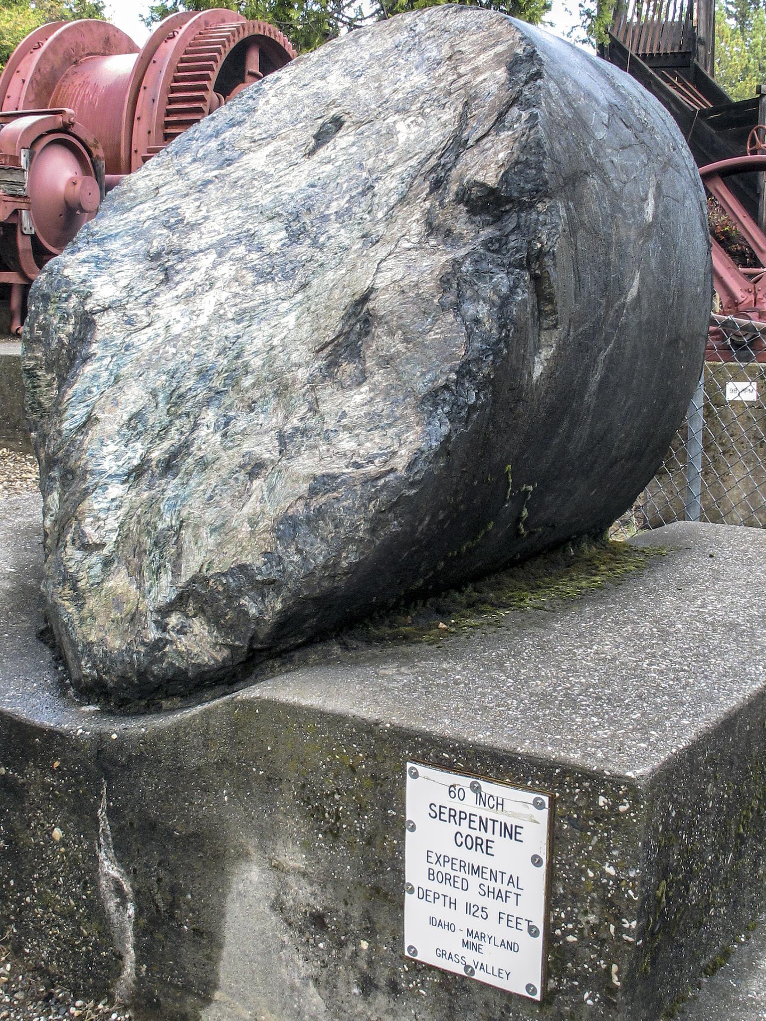 A large, dark, oblong rock sits on a concrete pedestal outdoors in daylight.