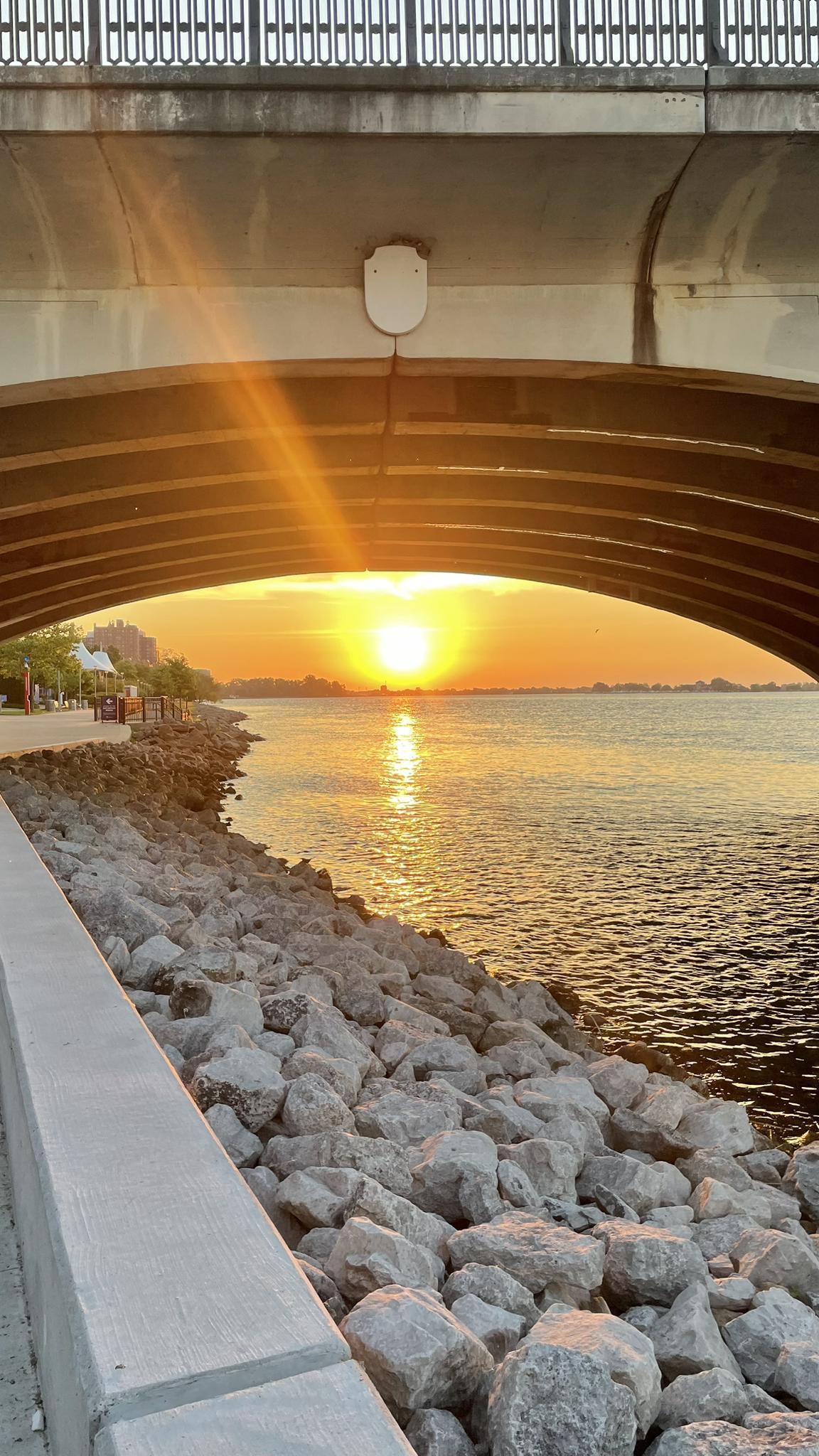 Golden sunset under a concrete bridge arch, reflecting on water and rocky shore.