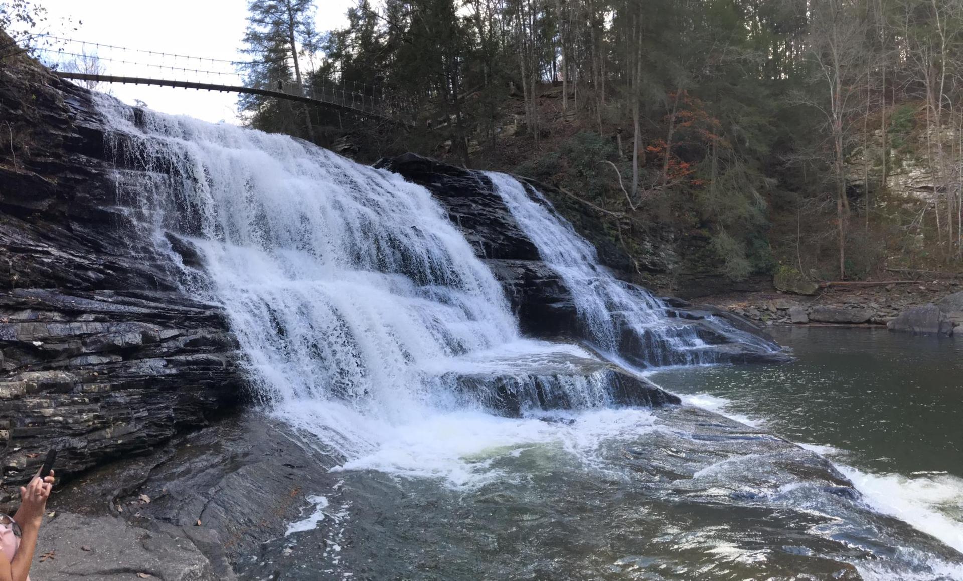 A wide waterfall cascades over dark rocks into a pool, with a bridge above.