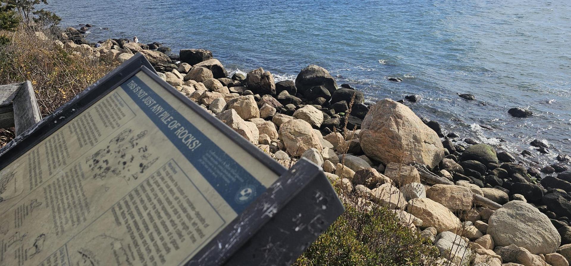 Interpretive sign overlooks a sunlit rocky coastline and calm blue ocean.