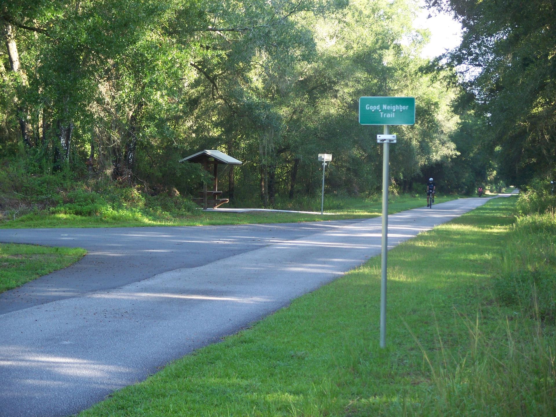 Sunny paved trail through woods with a cyclist and a sign.