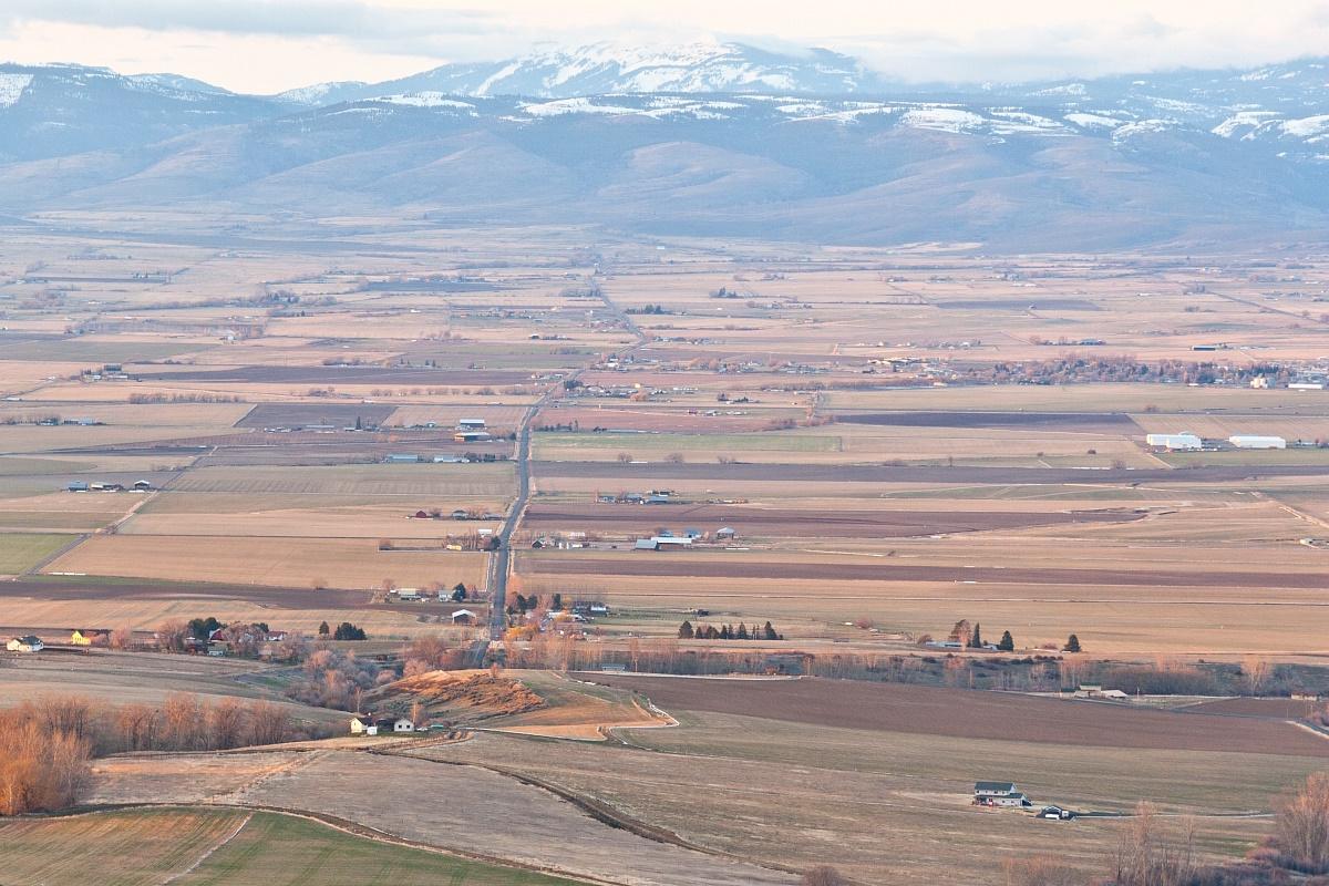 Vast valley with fields and a road, distant snow-capped mountains, soft light.
