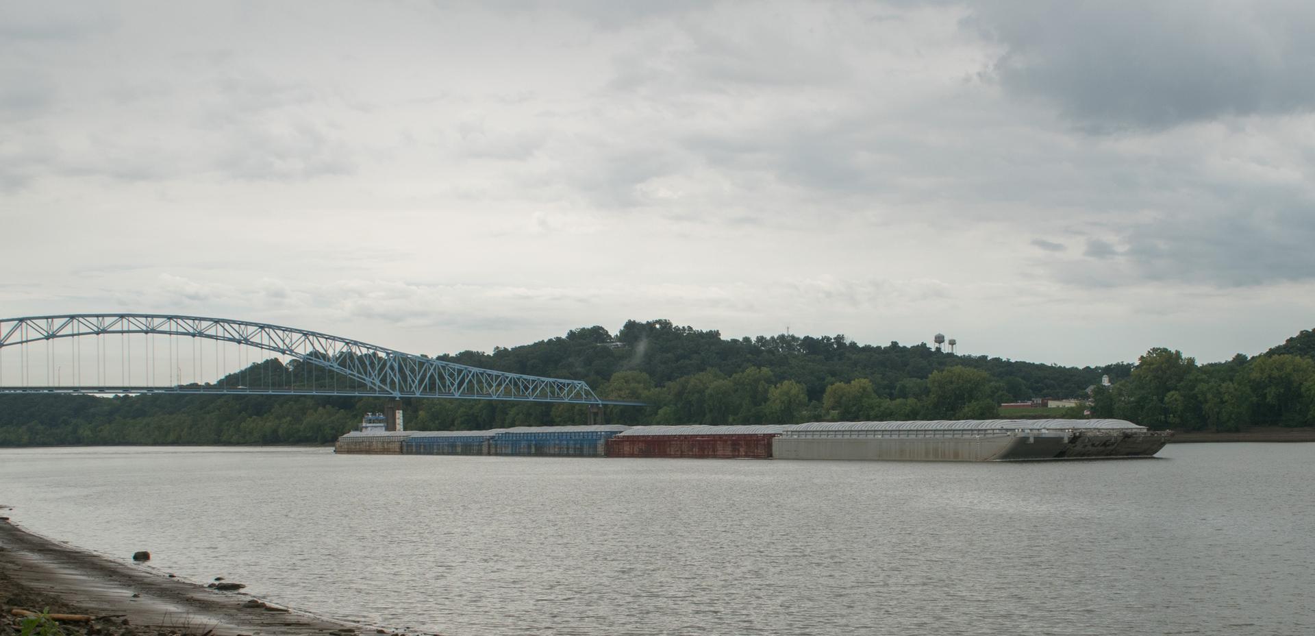 A large barge moves on a river, with a bridge and green hills, under a cloudy sky.