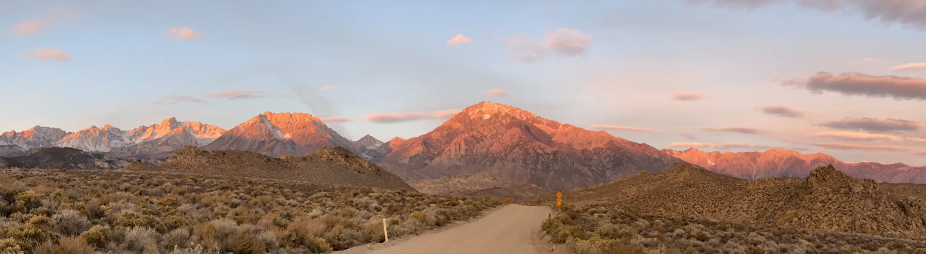 Dirt road leading to mountains bathed in warm sunset light with pink clouds.