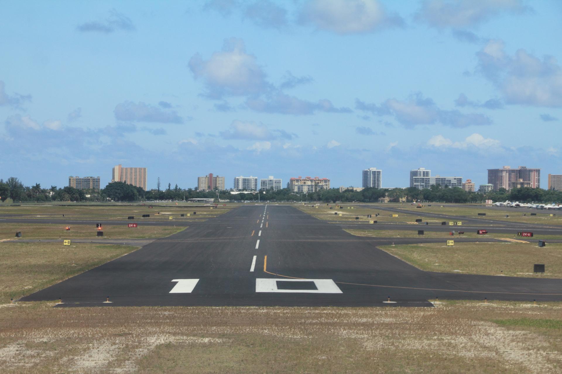 An empty airport runway stretches towards distant buildings under a partly cloudy sky.