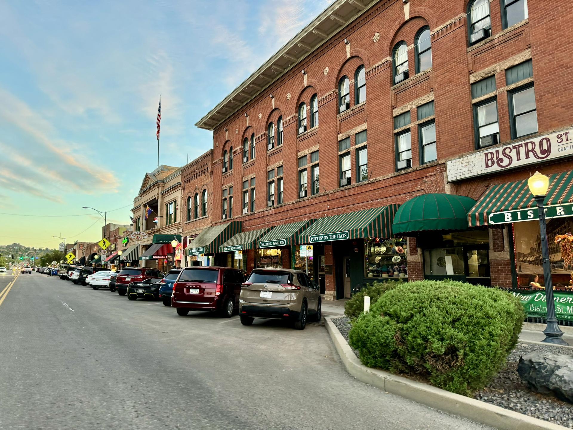 Urban street lined with brick buildings, parked cars, under evening light.