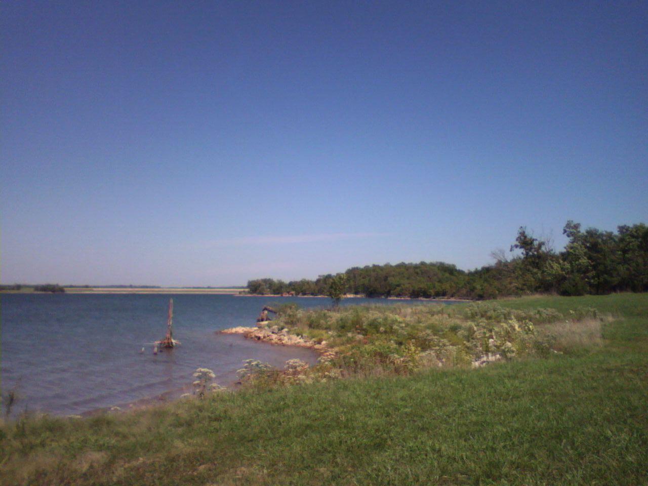 Calm lake under clear blue sky, with grassy and rocky shoreline.