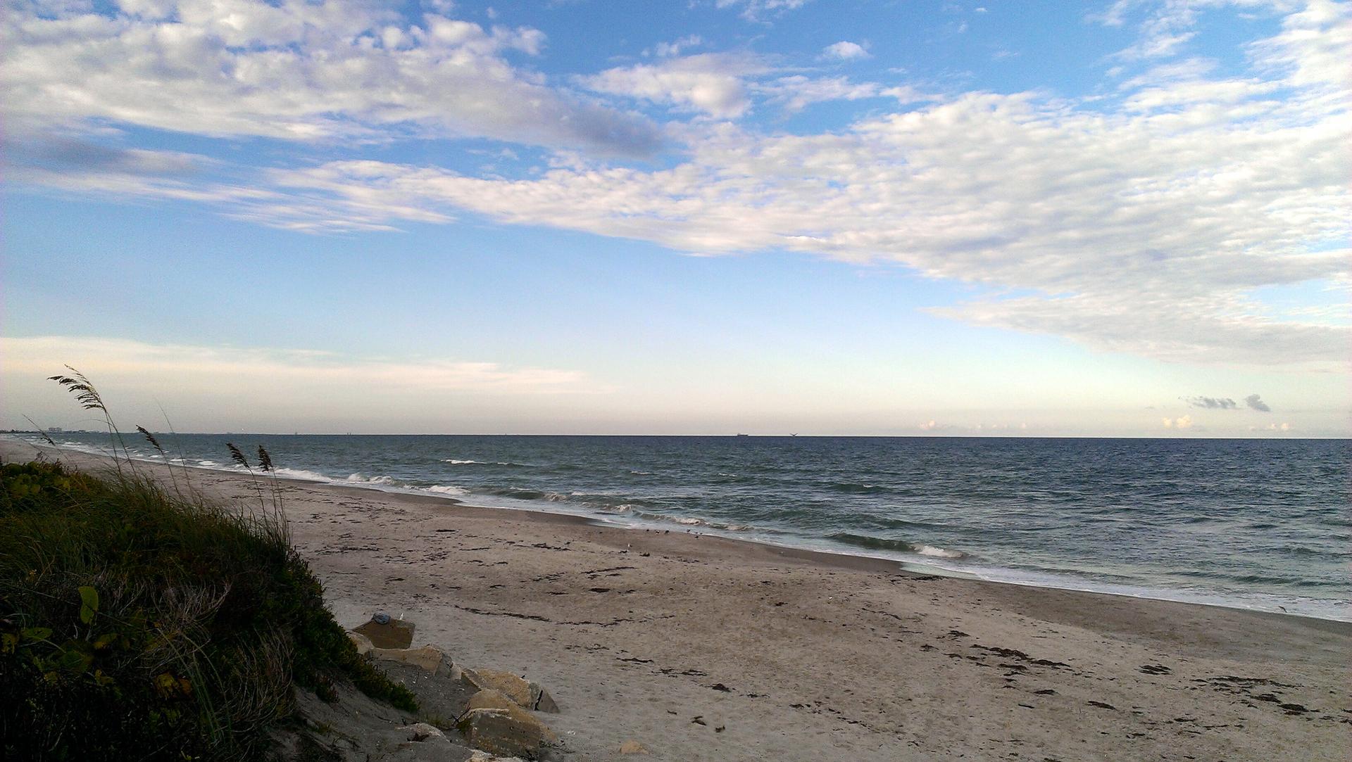 Wide sandy beach meets ocean waves under a partly cloudy blue sky.