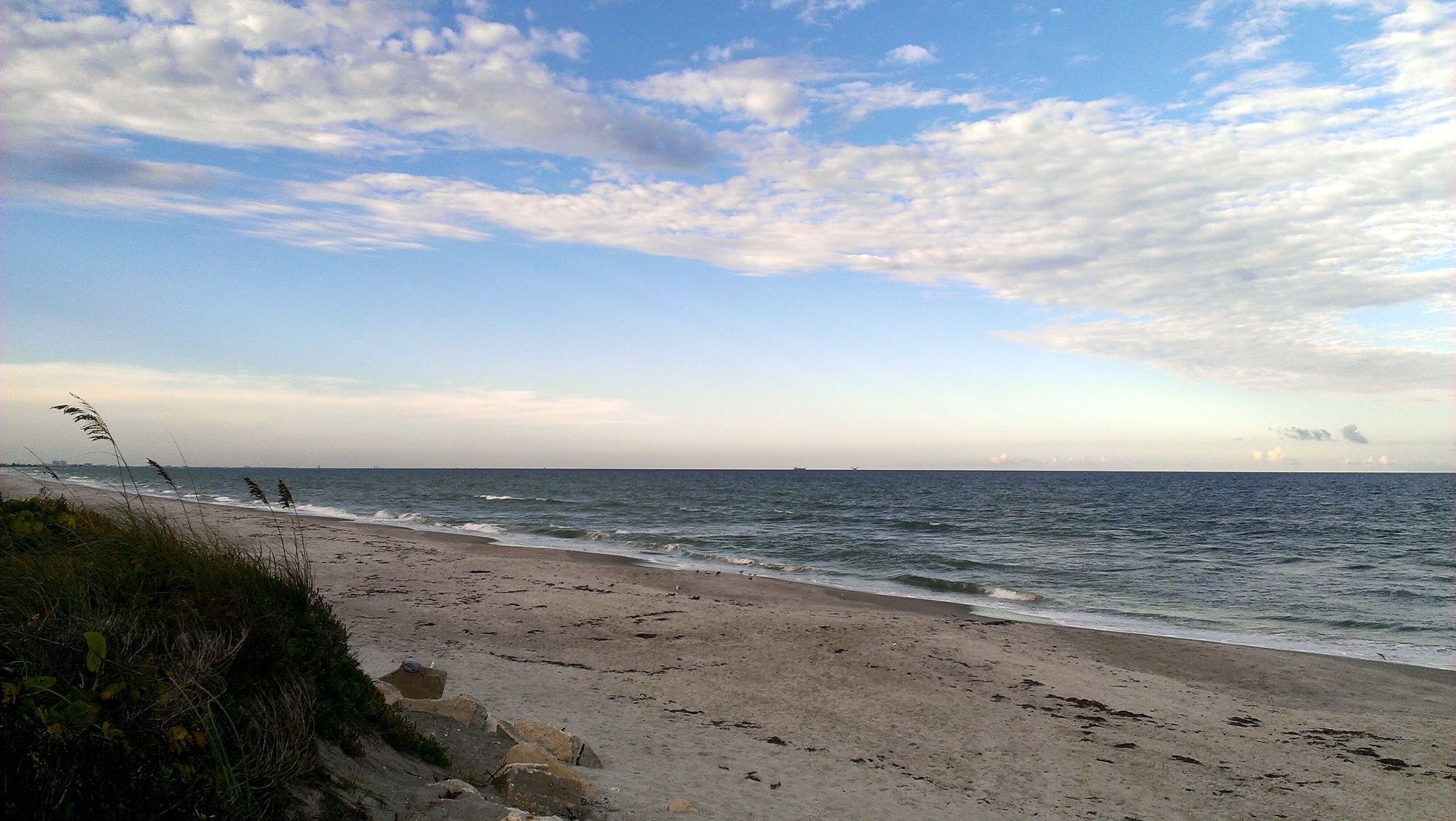 Wide sandy beach meets ocean waves under a partly cloudy blue sky. - Merritt Island loop via Satellite Beach
