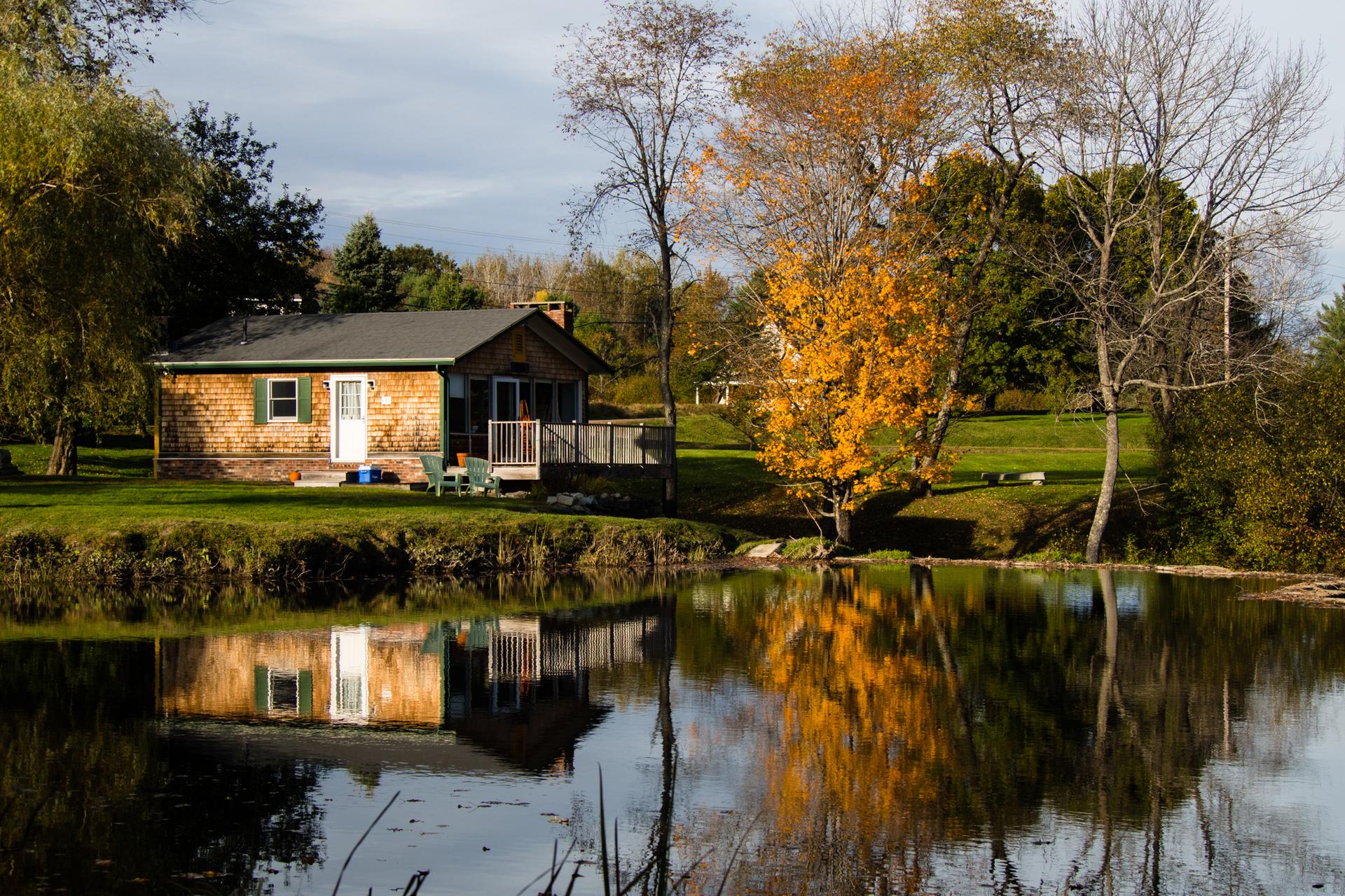 A cozy cabin by a calm pond, reflecting autumn trees under bright skies.
