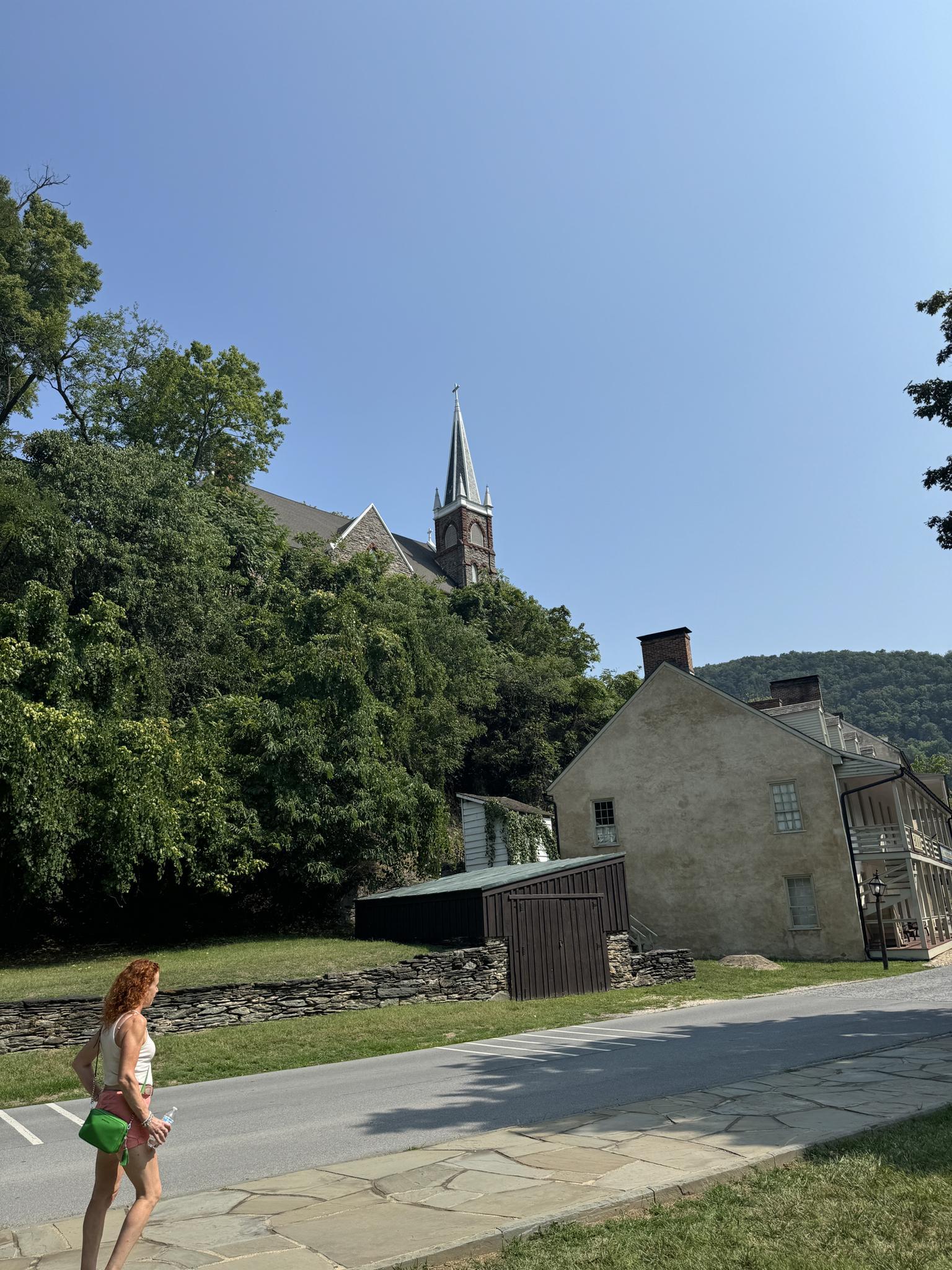 Woman walks on a sunny road past historic buildings and a church steeple.