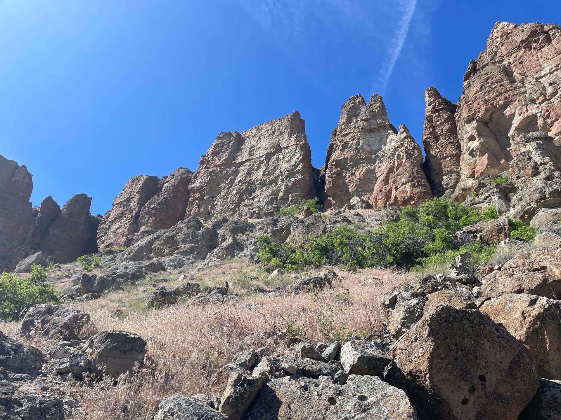 Towering, rugged rock formations rise from a dry, grassy hillside under blue sky.