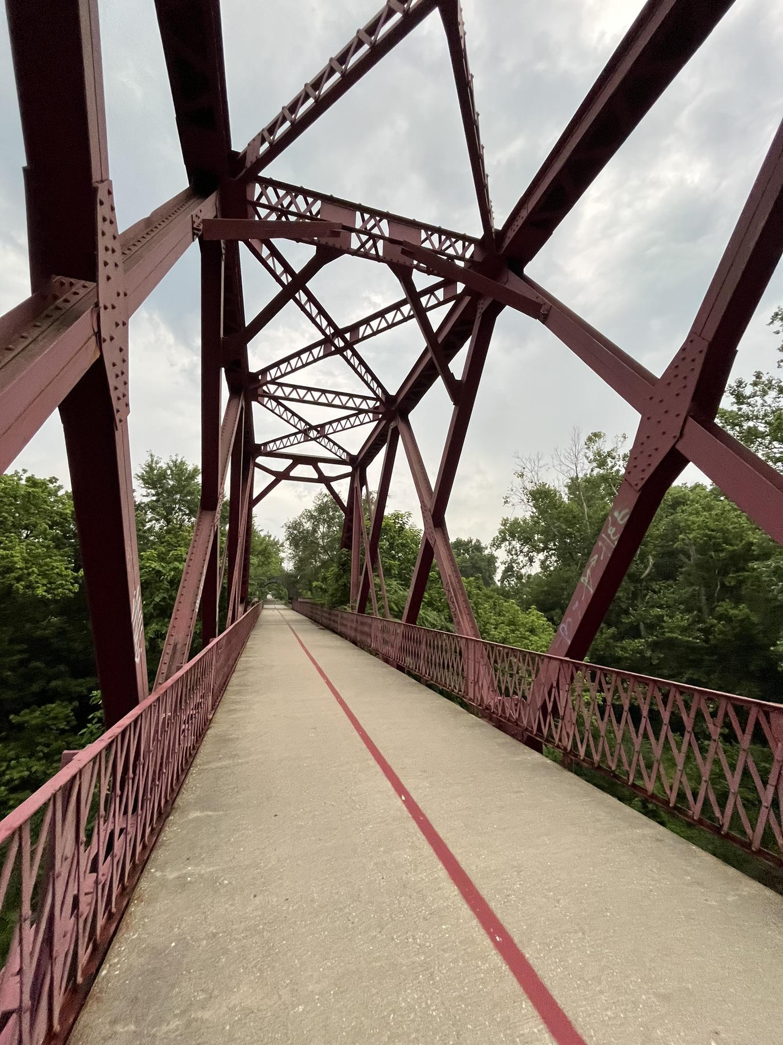 A red metal truss bridge with a paved path, surrounded by green trees.