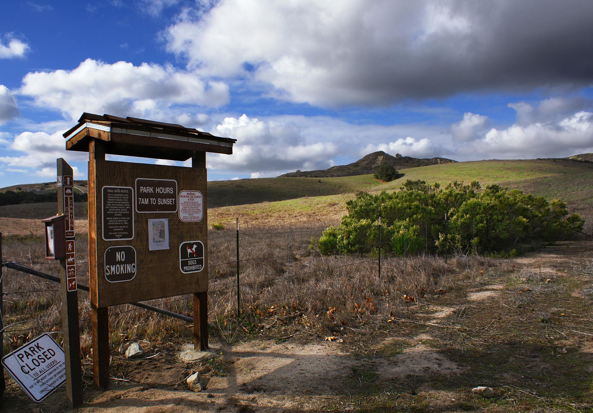 A wooden information sign and "PARK CLOSED" sign on a dirt path with dry hills.