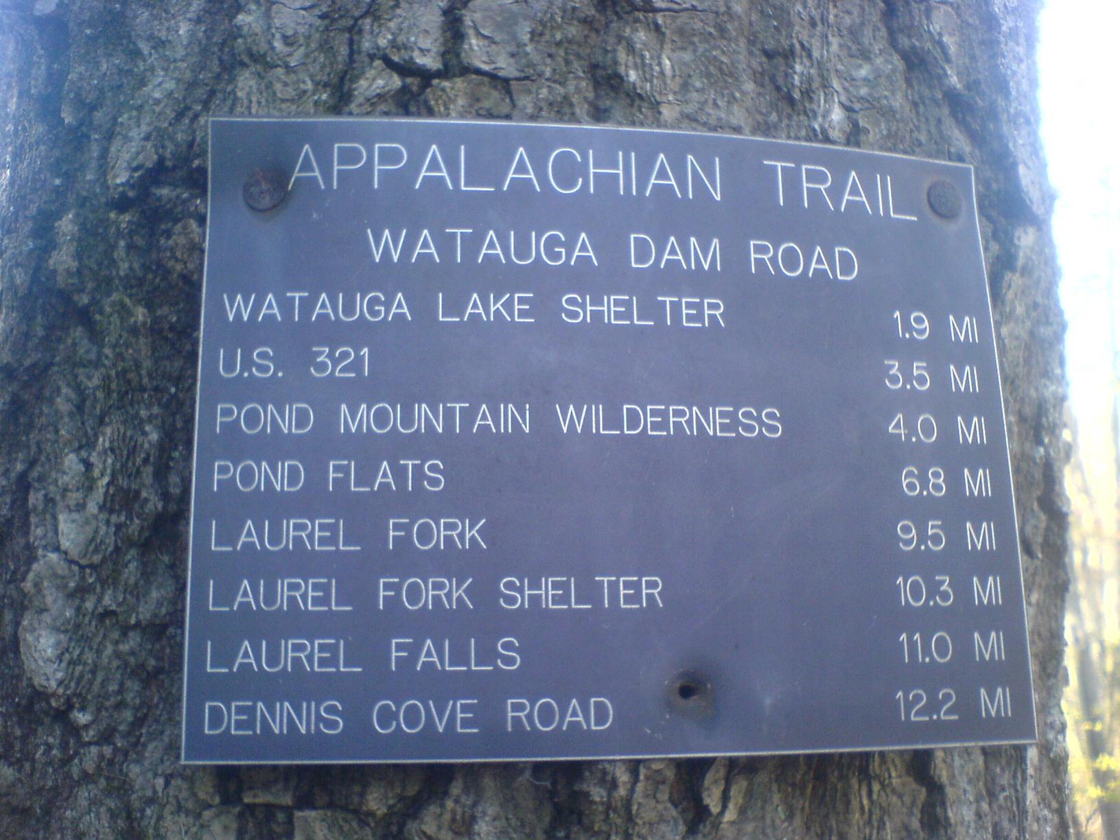 A metal trail sign with destination names and distances is affixed to a tree.