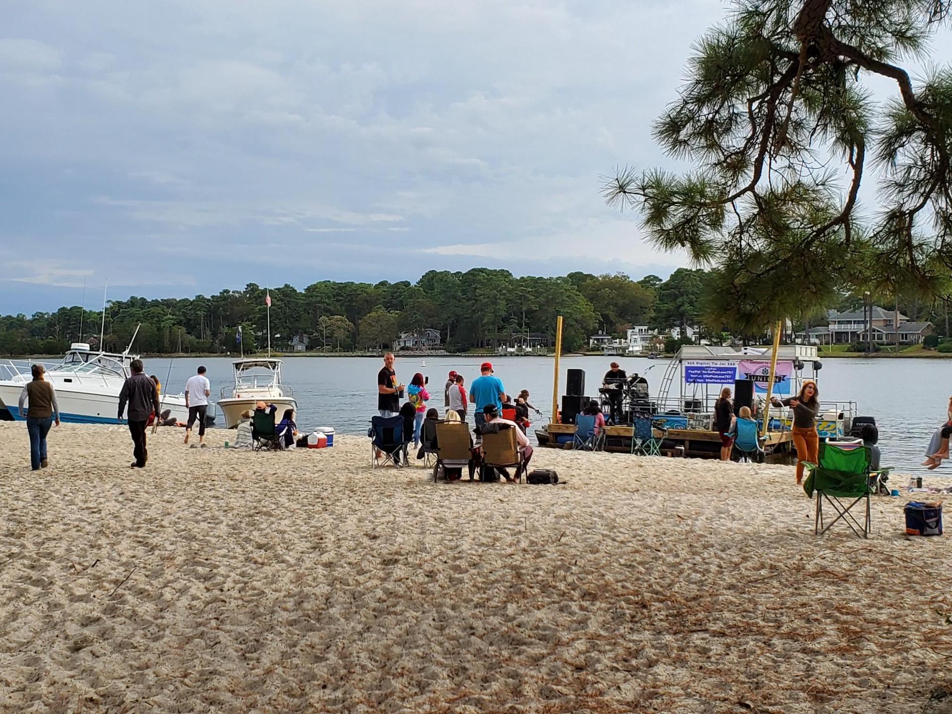 People gather on a sandy beach by a lake with boats and an outdoor stage.