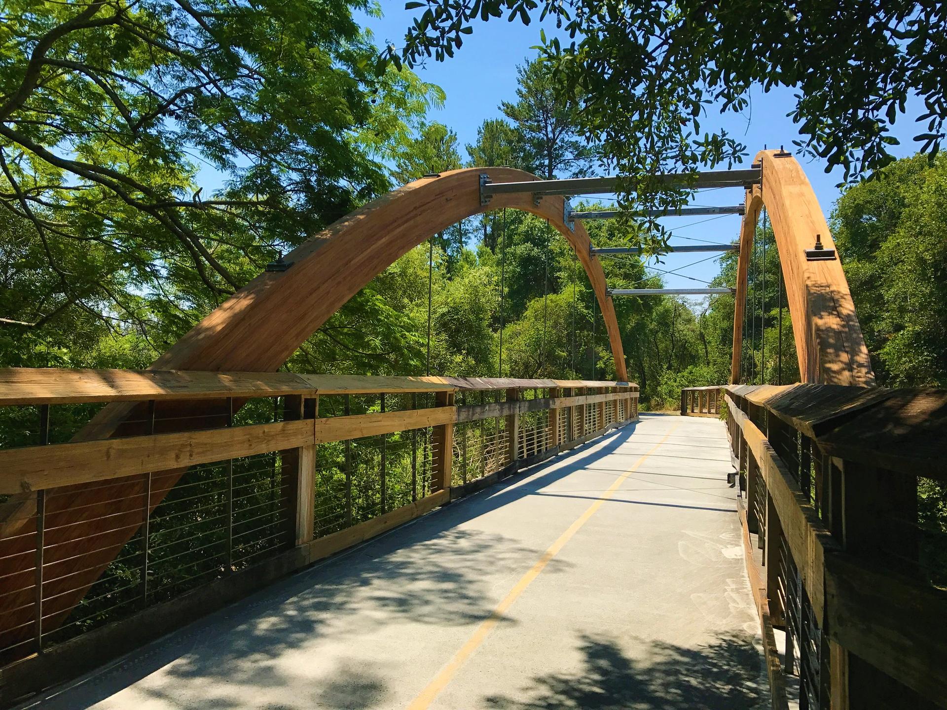A modern wooden arch bridge spans a paved path on a bright, sunny day.