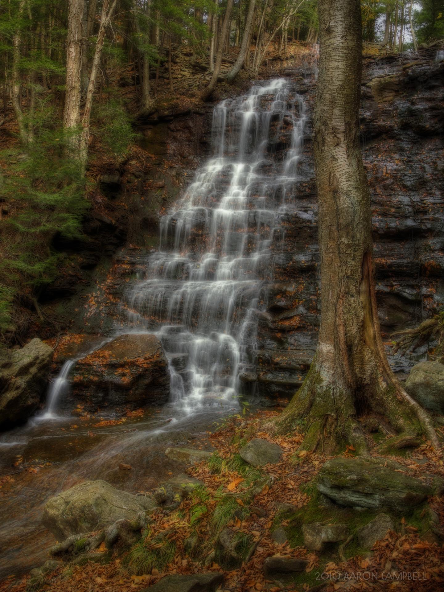 A multi-tiered waterfall cascades down a rocky cliff in an autumn forest.
