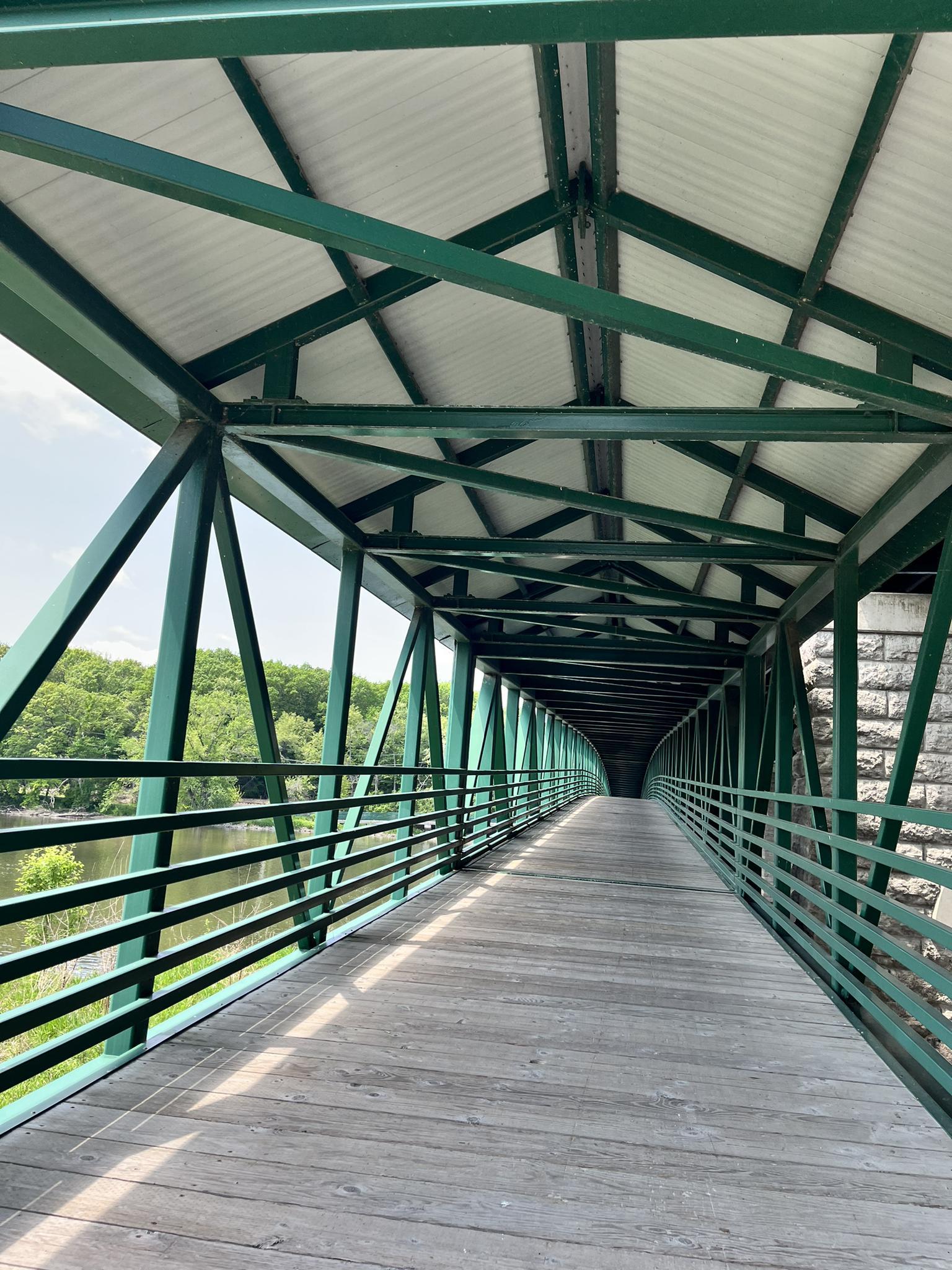 Long, green metal covered pedestrian bridge over water on a bright day.
