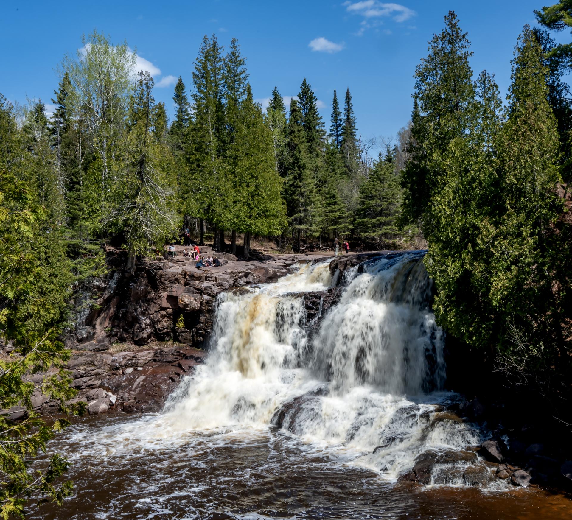 People gather near a powerful waterfall surrounded by green forest on a sunny day.