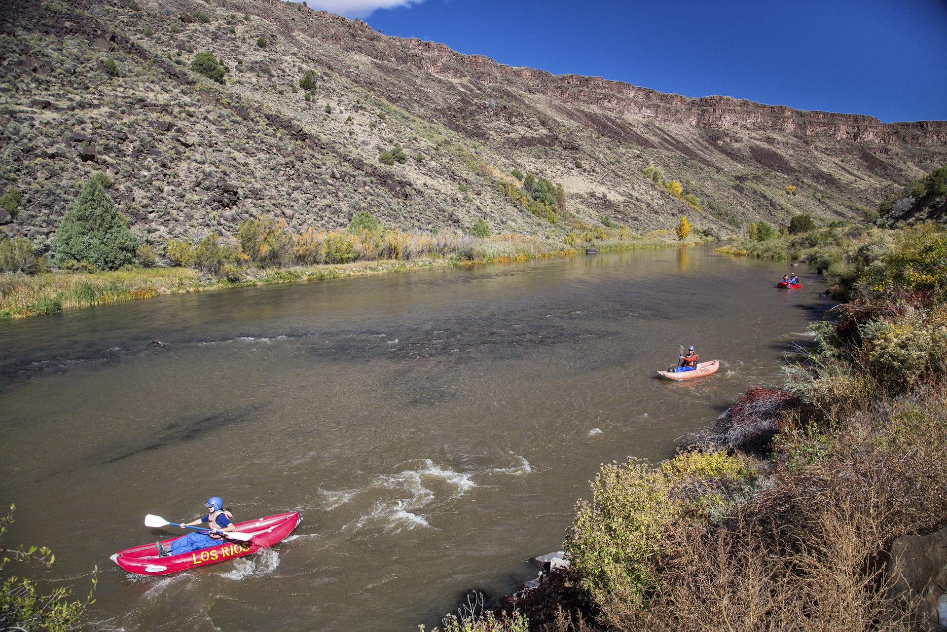 Two kayakers paddle on a river through a rocky canyon on a sunny day.