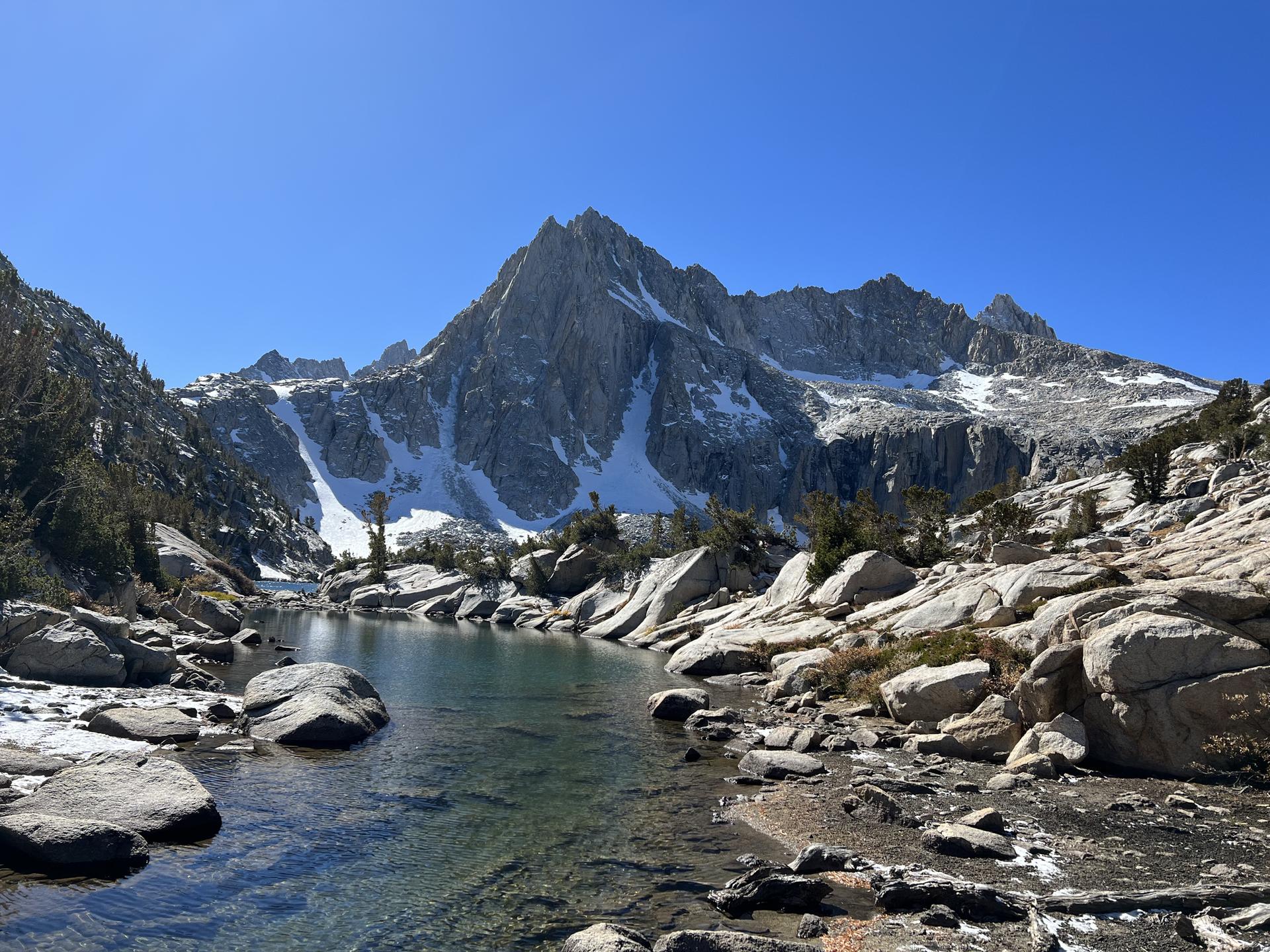 Clear mountain stream flowing into a lake, with snow-capped peaks and rocks.