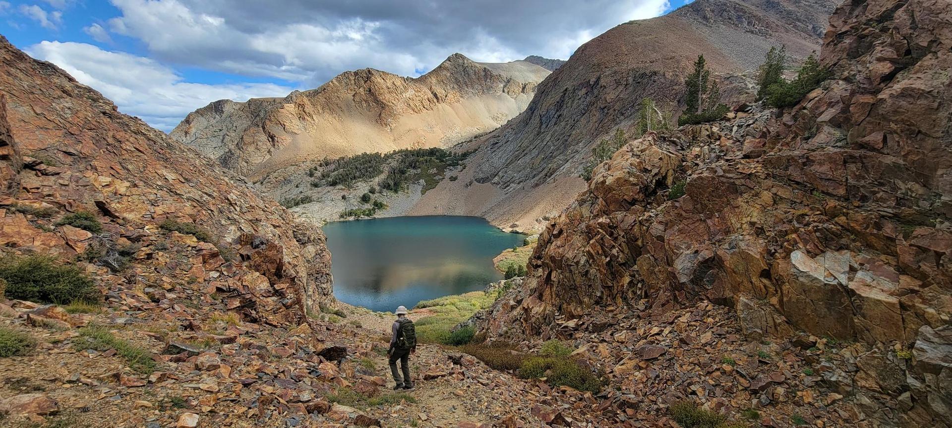Hiker views a turquoise mountain lake surrounded by rocky slopes under cloudy sky.