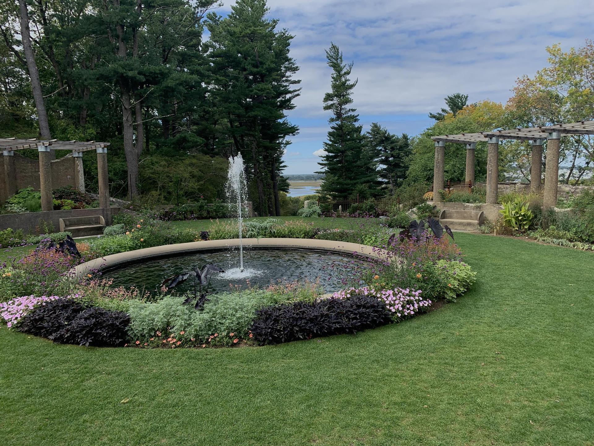 A circular fountain surrounded by flowers and green lawn under a cloudy sky.