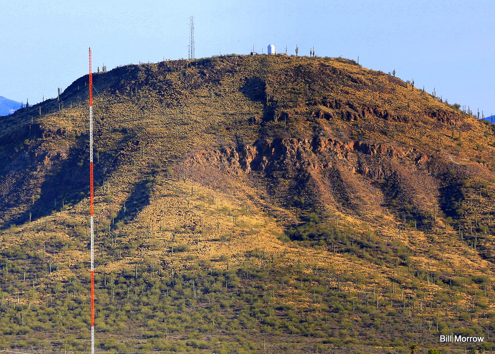 Large, dry desert mountain covered in sparse vegetation and cacti, with a tall antenna.