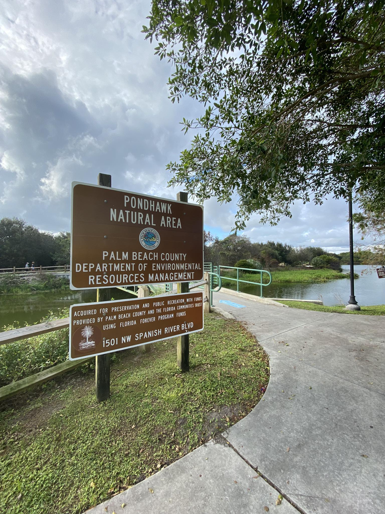 A brown sign stands beside a paved path and calm water on a cloudy day.