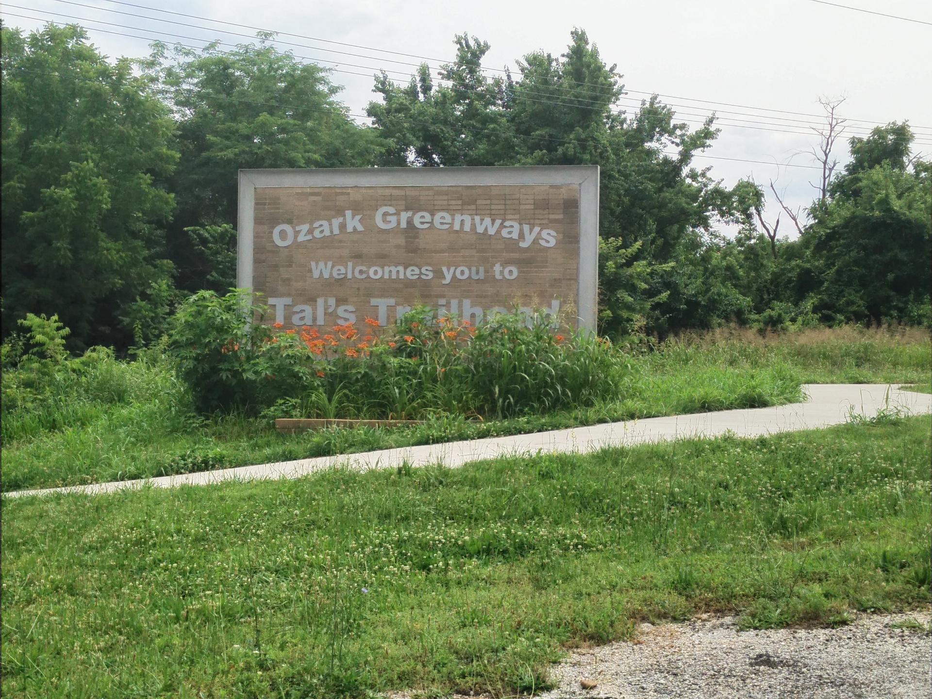 A large welcome sign stands amidst green foliage beside a paved path.