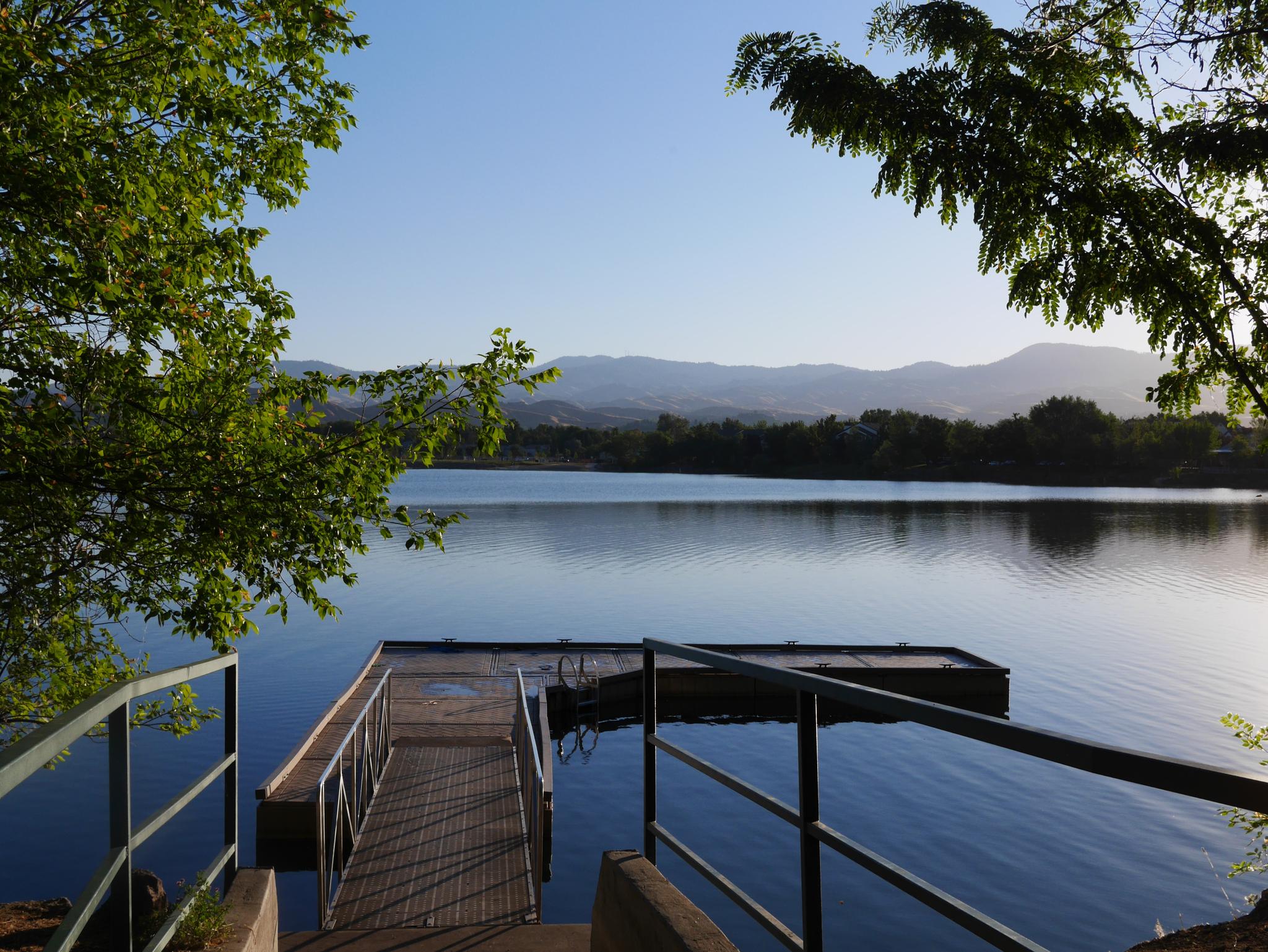 Wooden dock on a calm lake, framed by trees, with distant mountains.