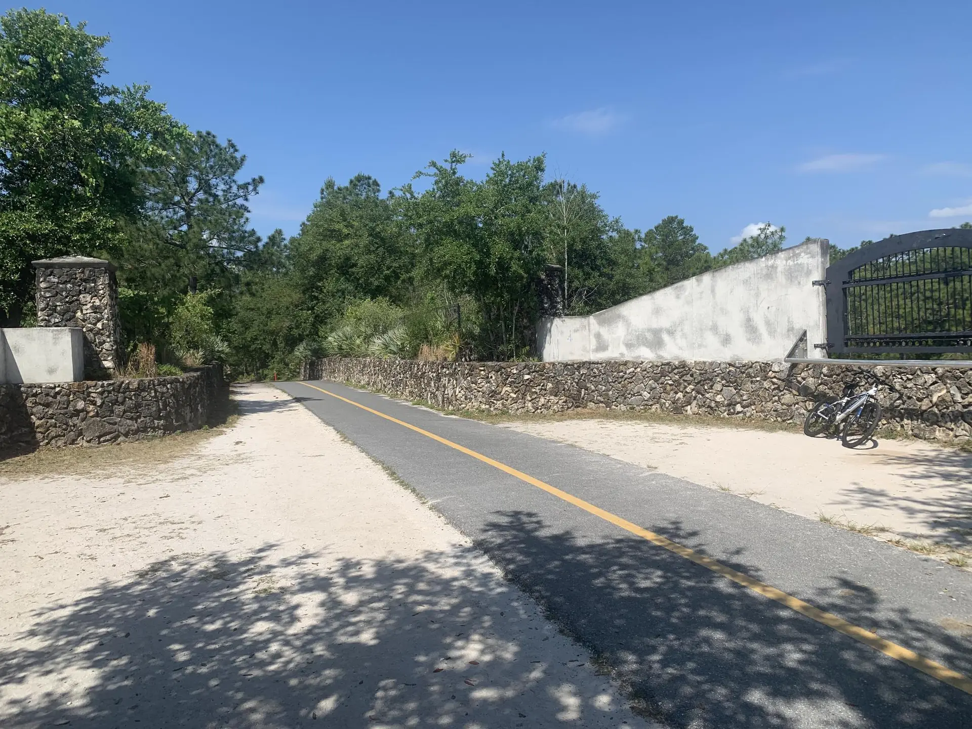 A paved road with stone walls and a gate under a bright, sunny sky. A bicycle is parked.