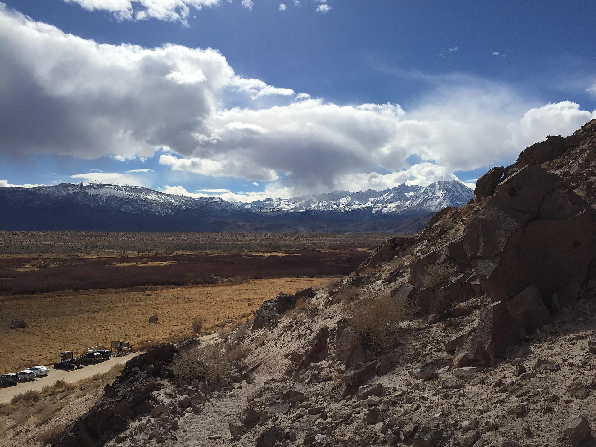 Snow-capped mountains overlook a wide valley with parked cars under a cloudy sky.