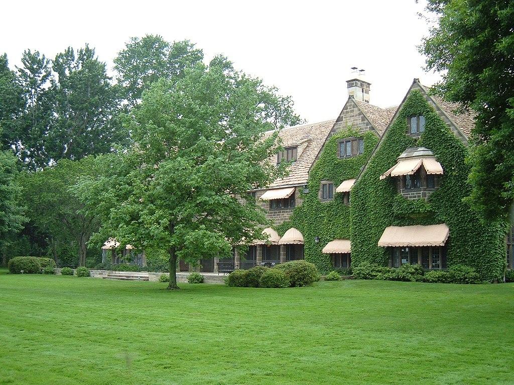 A large, ivy-covered house with awnings stands behind a vibrant green lawn and trees.