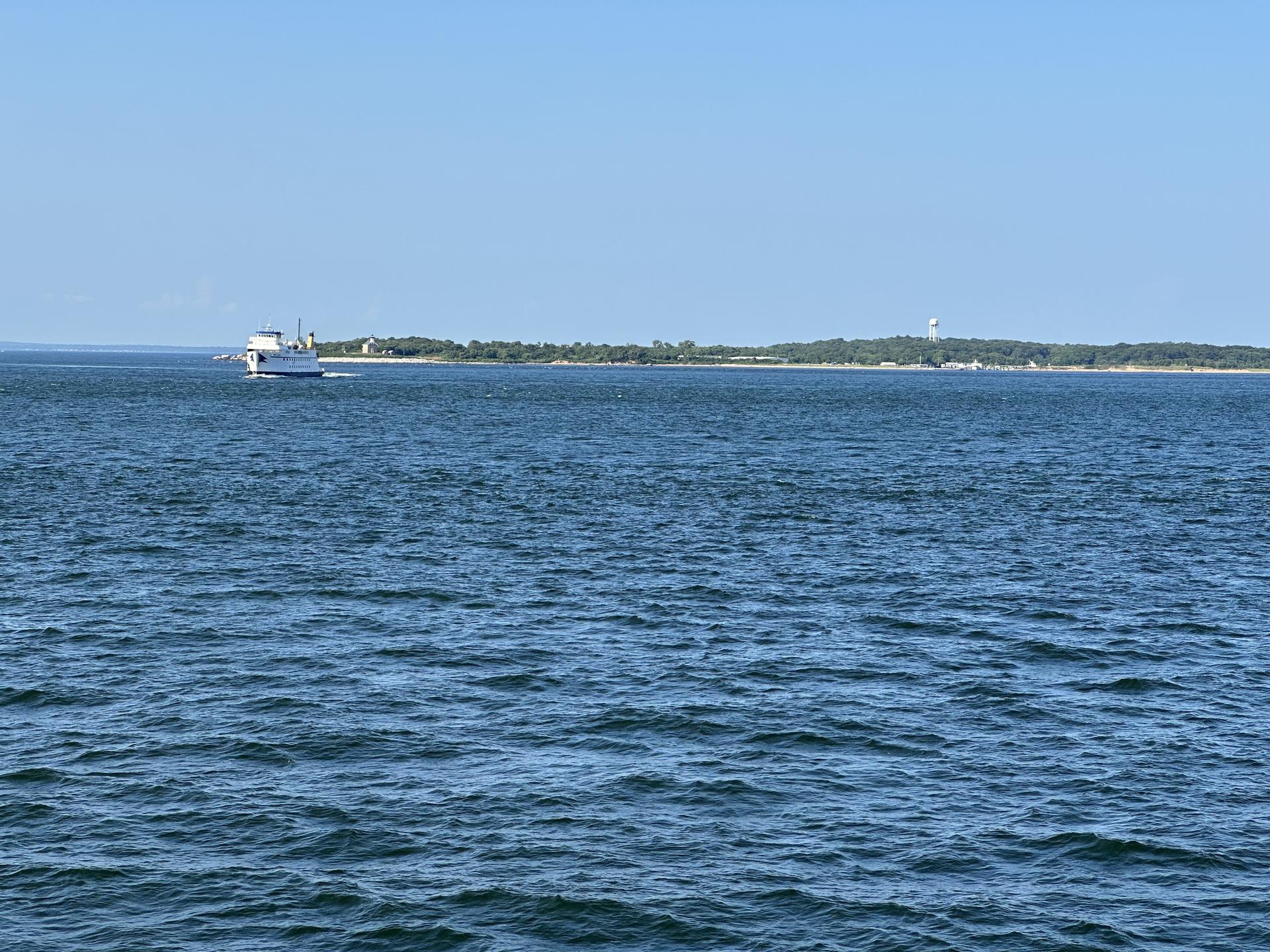 A boat crosses calm blue water towards a distant tree-lined shore under a clear sky.