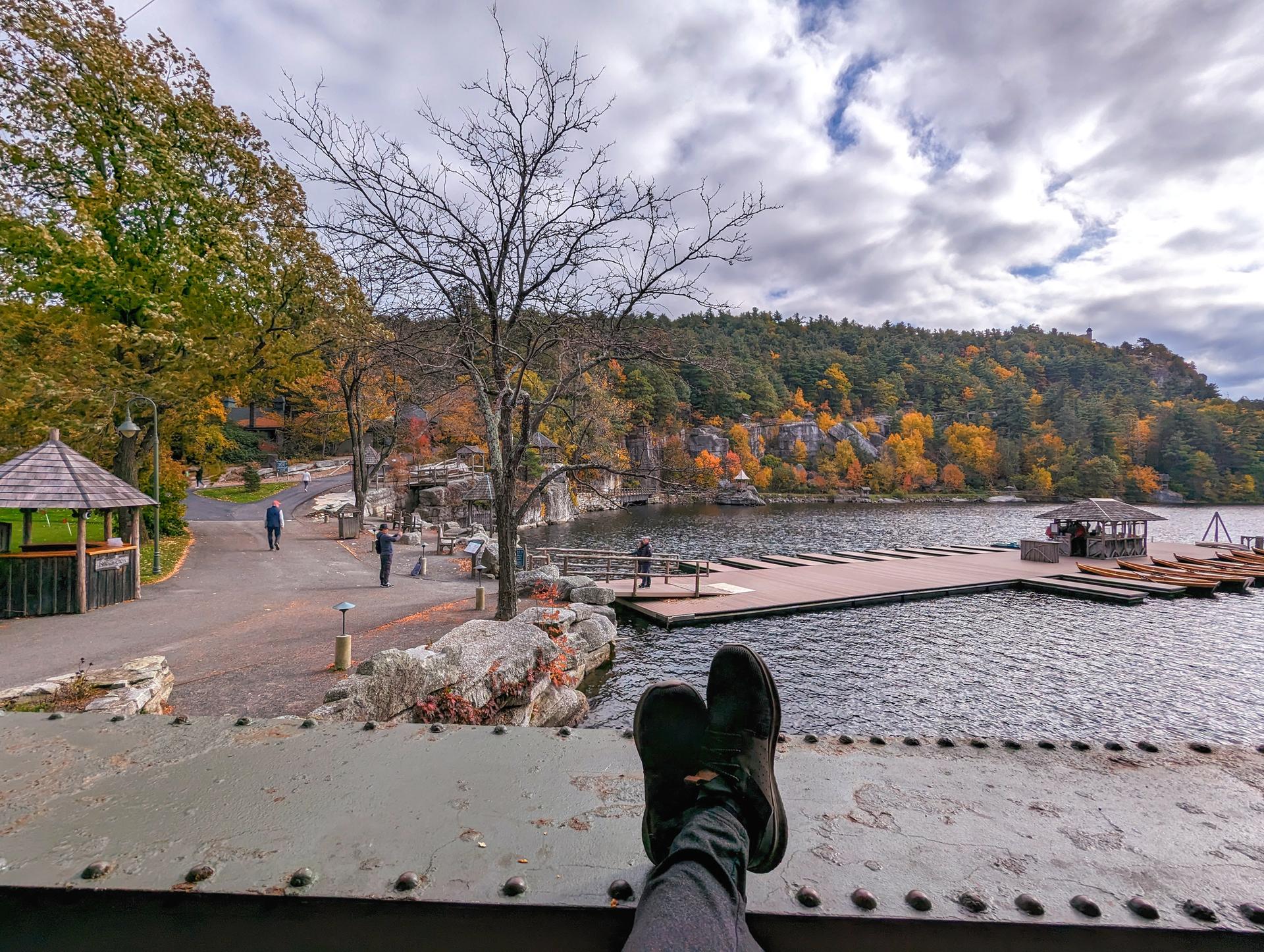 Person's feet resting, overlooking a lake, dock, and colorful autumn trees.