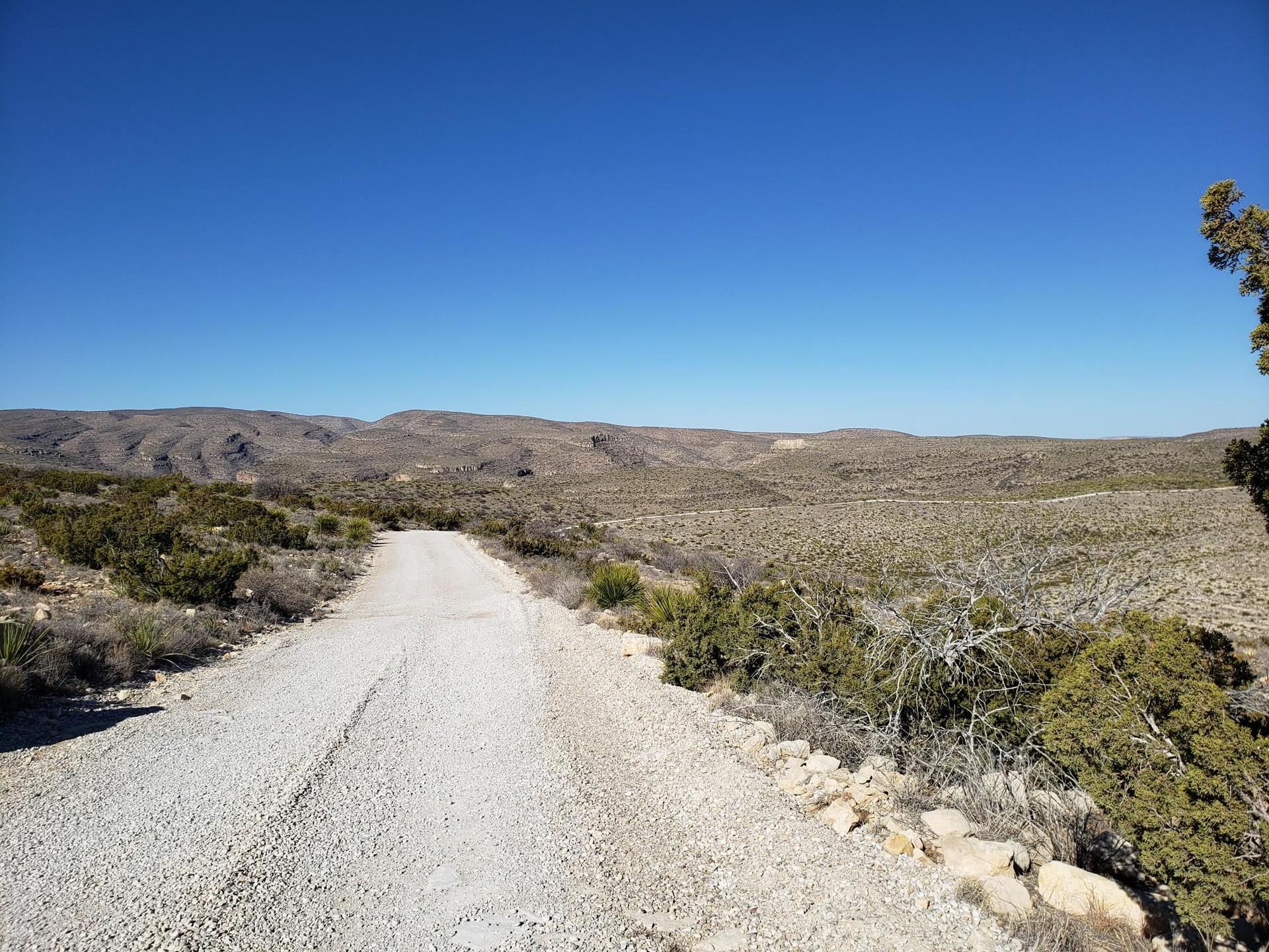 A gravel road stretches through a vast, sunny desert landscape with distant hills.