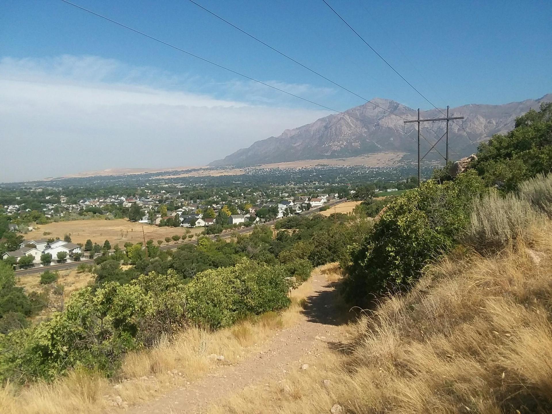 Dirt path on a hillside with a distant town and large mountain under blue sky.